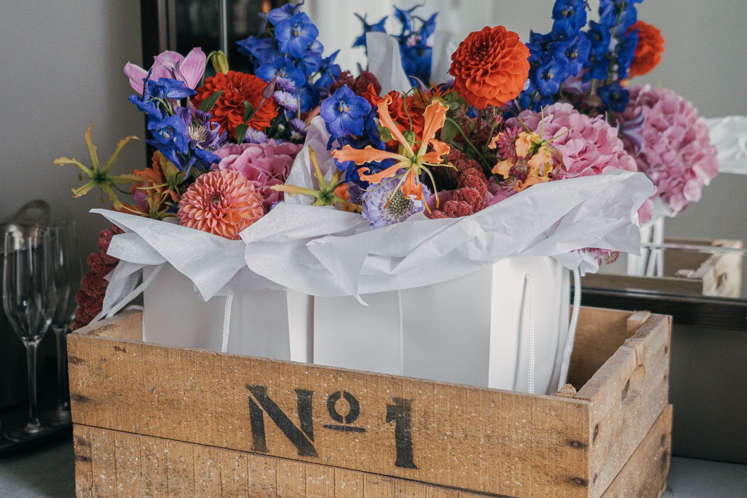 A rustic wooden crate marked 'No 1' holds a vibrant arrangement of blue, orange, and pink flowers wrapped in white paper.