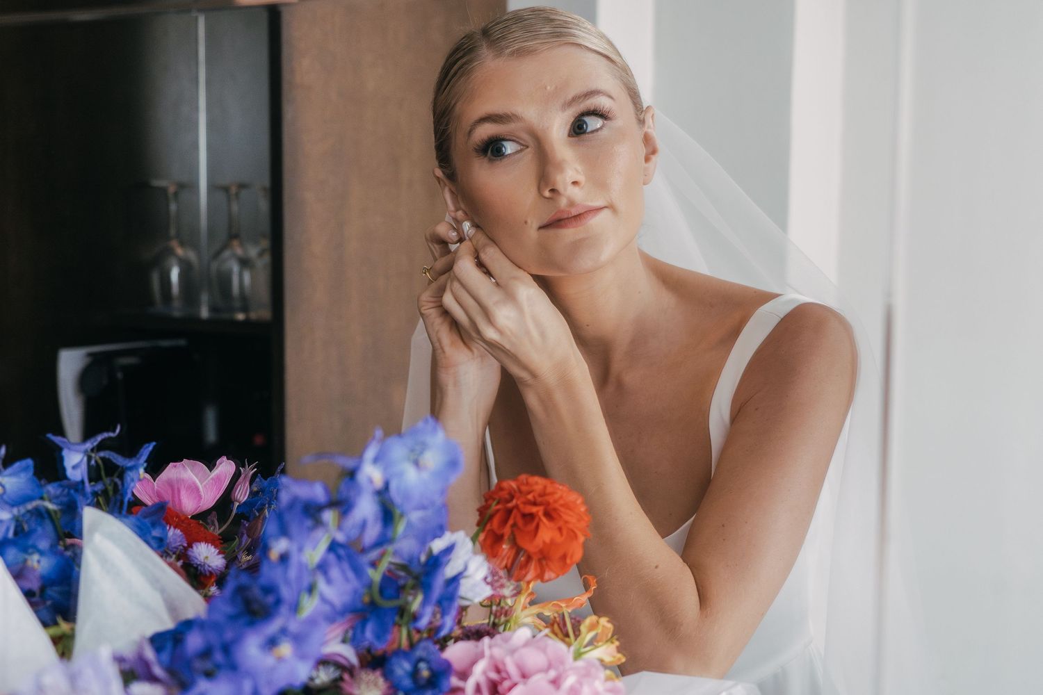 A bride looking thoughtful at a window with colorful blue and pink flowers in the foreground.