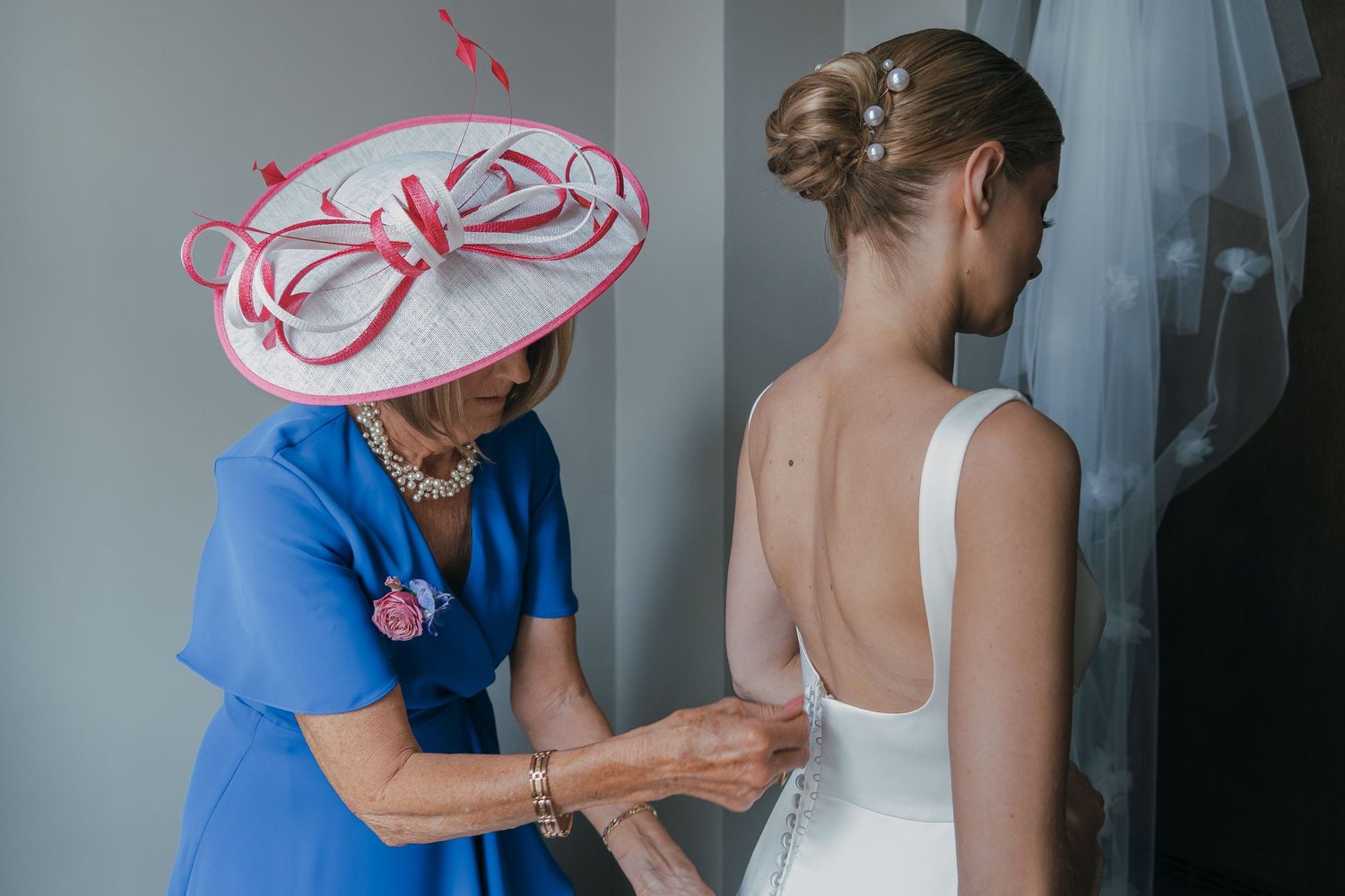 A wedding dress being carefully buttoned up with a person in a bright blue dress and pink hat assisting.