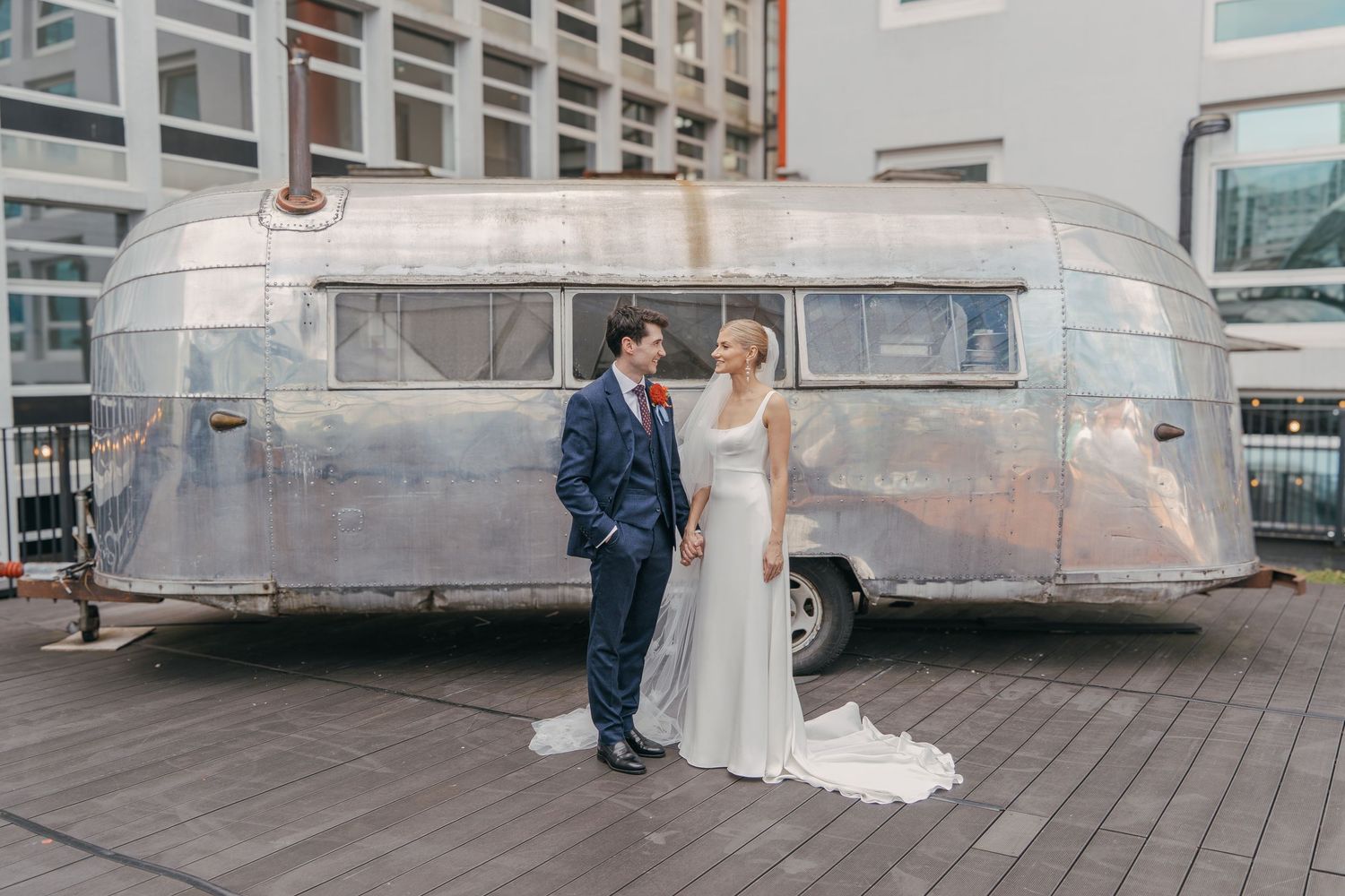 A couple poses together in front of vintage Airstream trailers on a wooden deck.