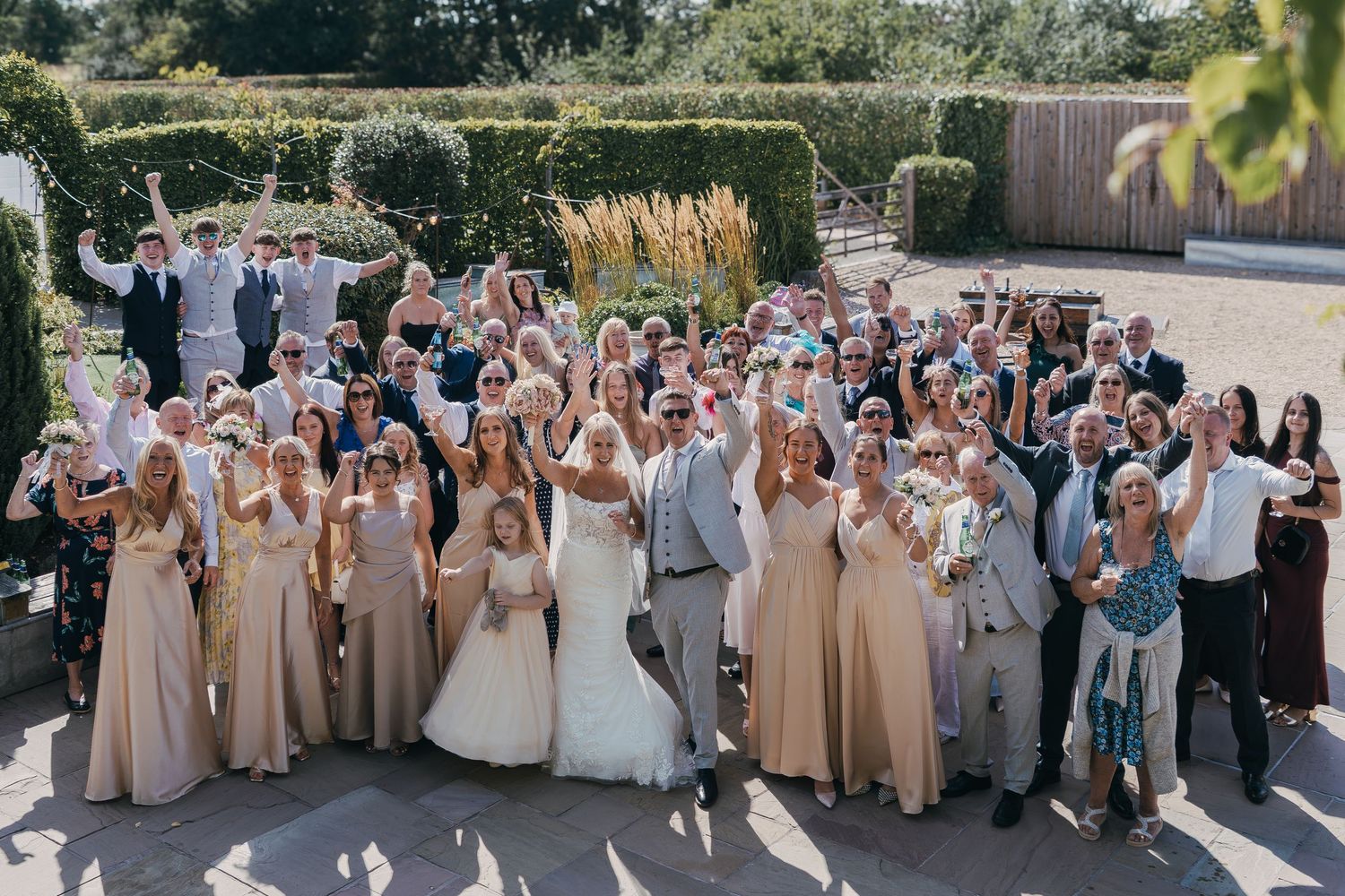 Wedding party group poses together in beige bridesmaids dresses and formal attire during outdoor celebration.