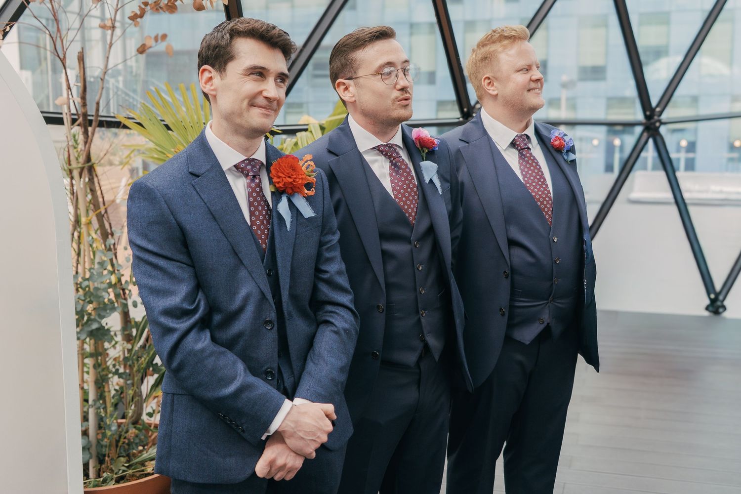 Three men in formal suits and red boutonnieres stand together at a wedding ceremony in a modern glass-walled venue.