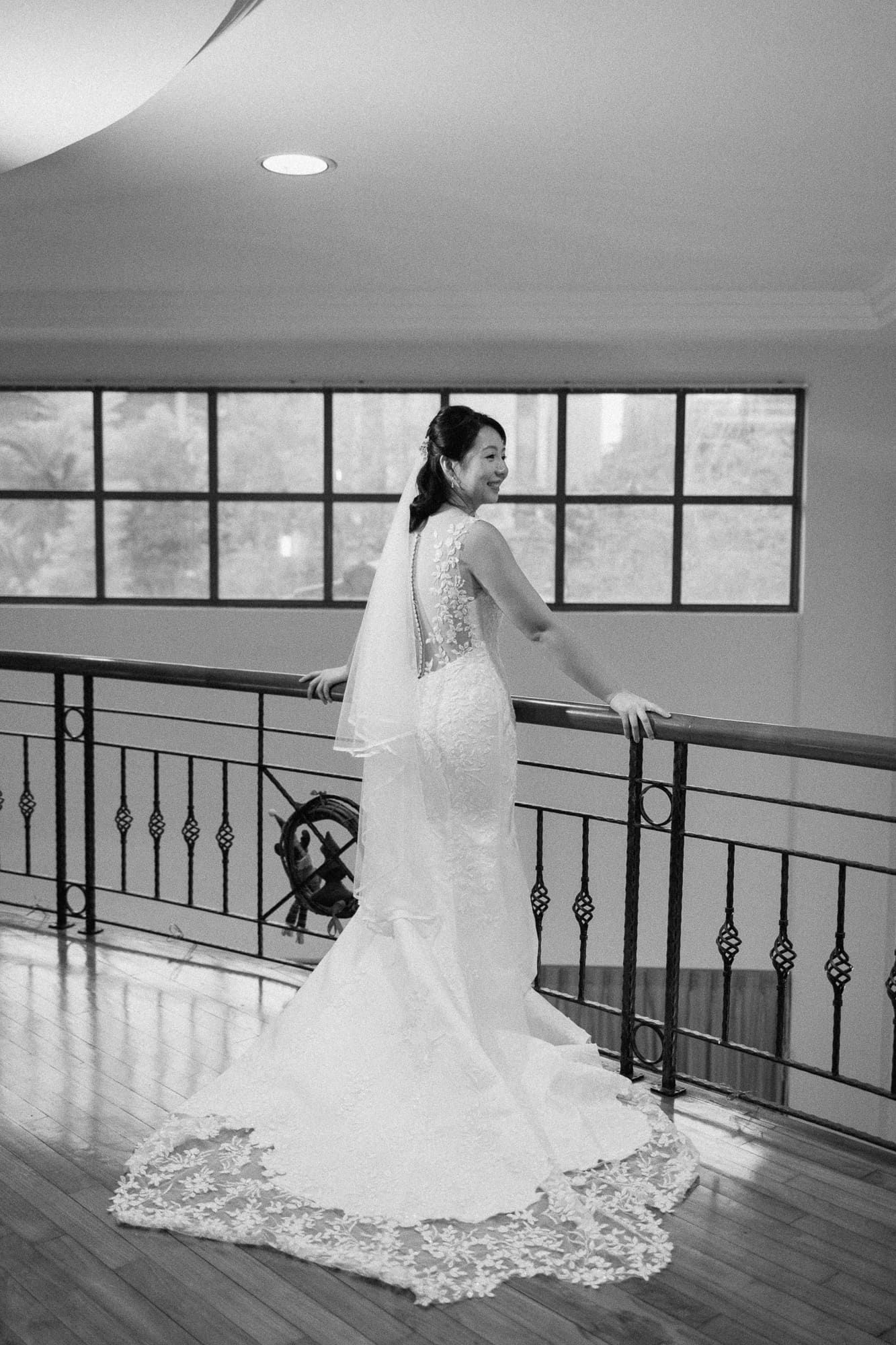 A bride in a white mermaid wedding dress sits gracefully on hardwood floor stairs with black metal railings.