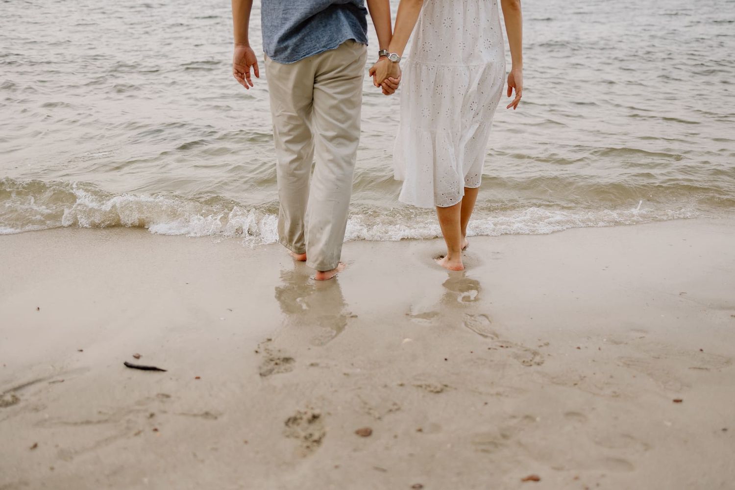 Legs of a couple standing in shallow ocean waves on a sandy beach.