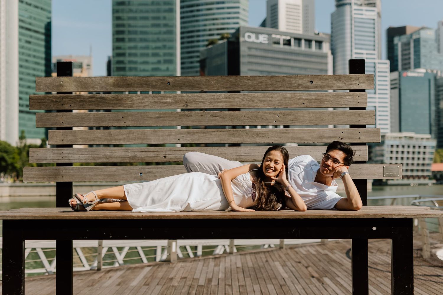 A playful couple lies down on a wooden bench overlooking a cityscape of tall buildings.