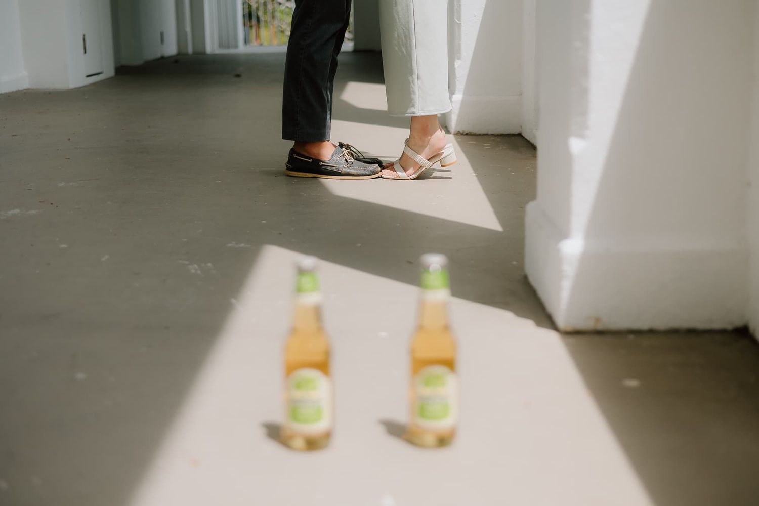 Two glass bottles casting shadows on white floor near standing feet.