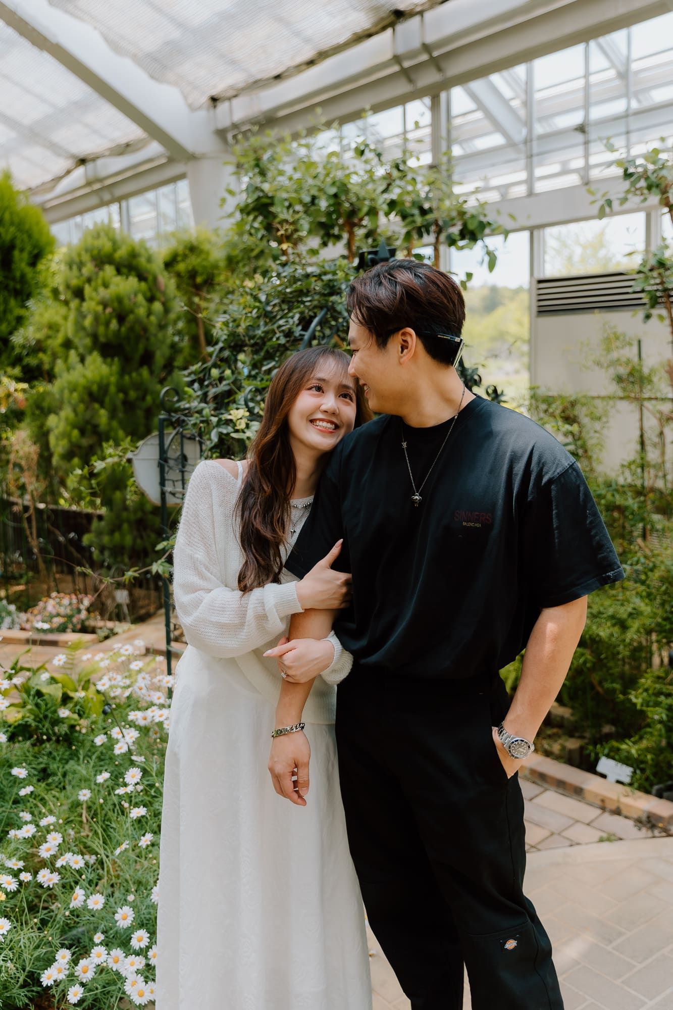 A couple embracing and smiling in a lush greenhouse, woman in white dress, man in black outfit.