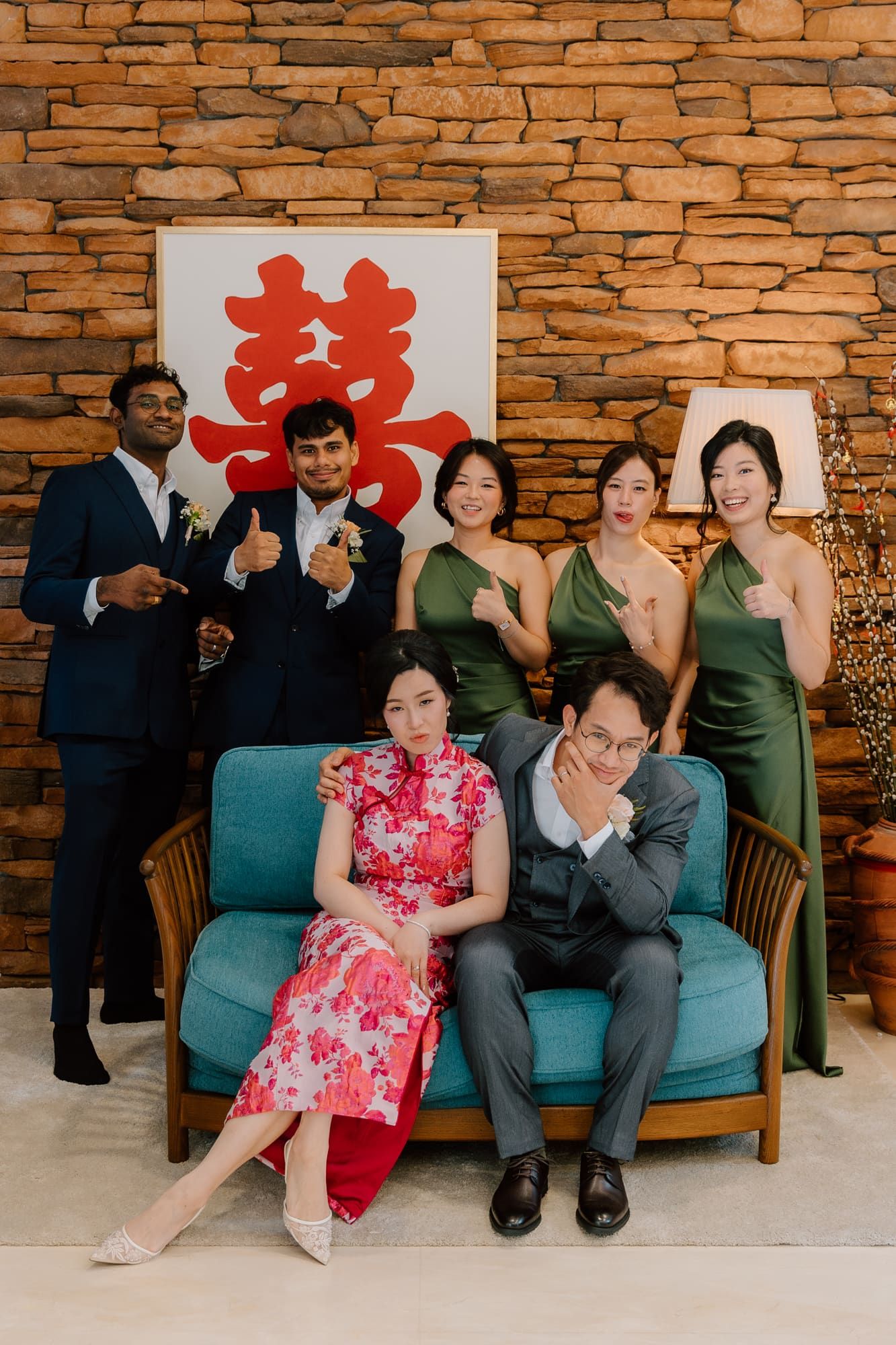 Wedding couple and guests pose playfully on a blue couch against a rustic stone wall with red double happiness symbols.