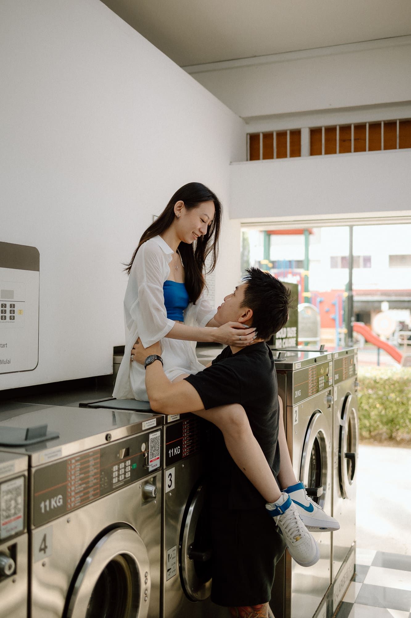 Two people embrace romantically in a laundromat setting with washing machines visible in the background.