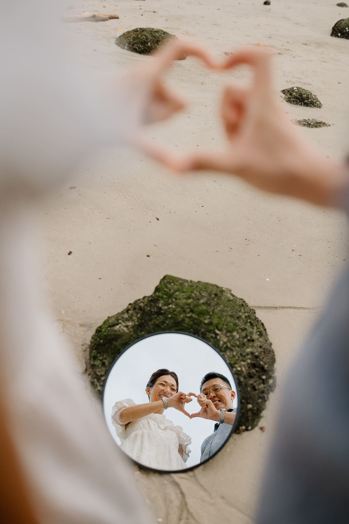 Creative wedding photo showing a couple making heart shapes with their hands reflected in a mirror on the beach.