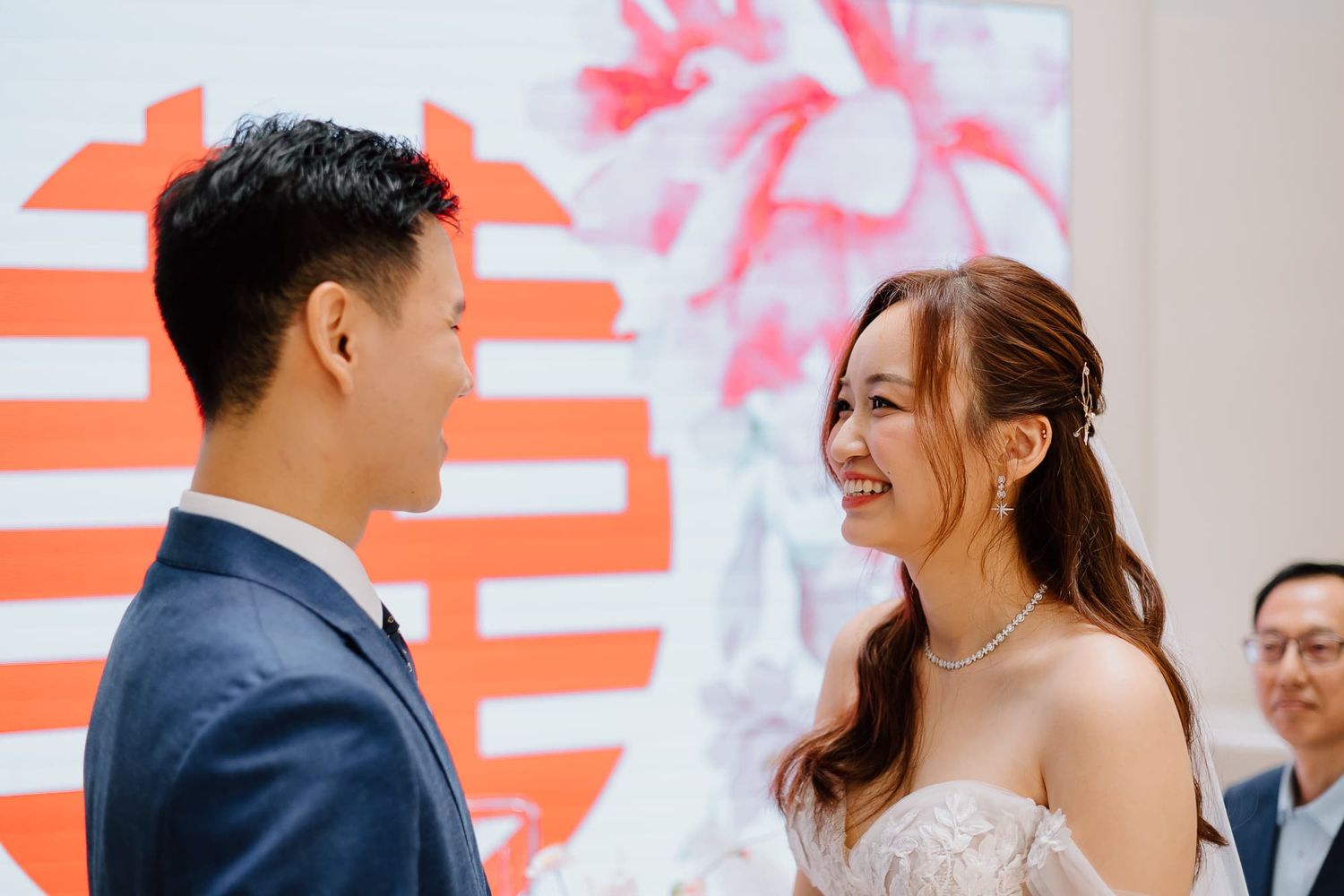 Couple shares a joyful moment in front of a decorative wall with pink floral designs and orange Chinese characters.