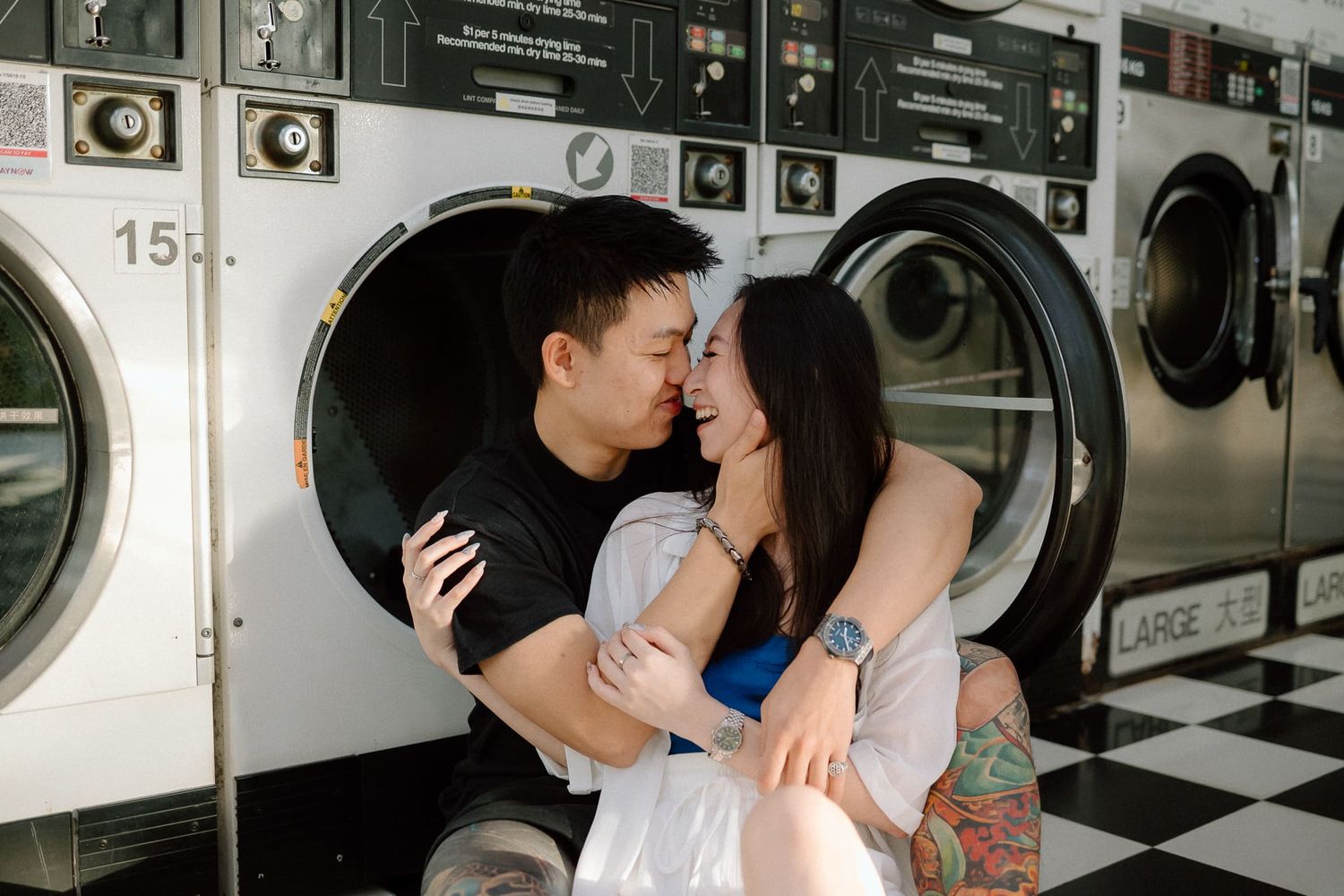 A romantic couple embraces and kisses while doing laundry at a retro-styled laundromat with black and white checkered floors.