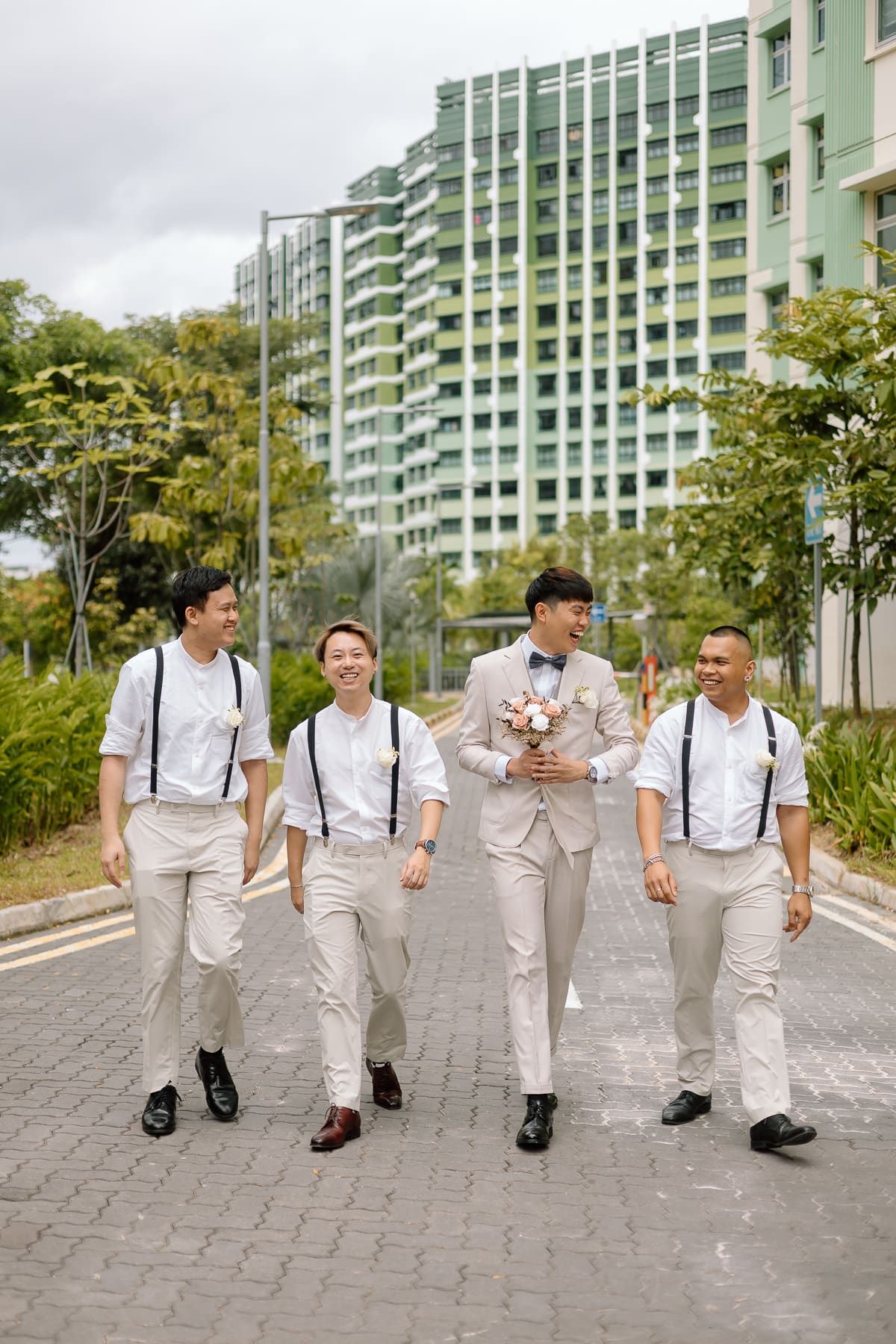 Groomsmen in matching white outfits and suspenders walking together along a pathway.