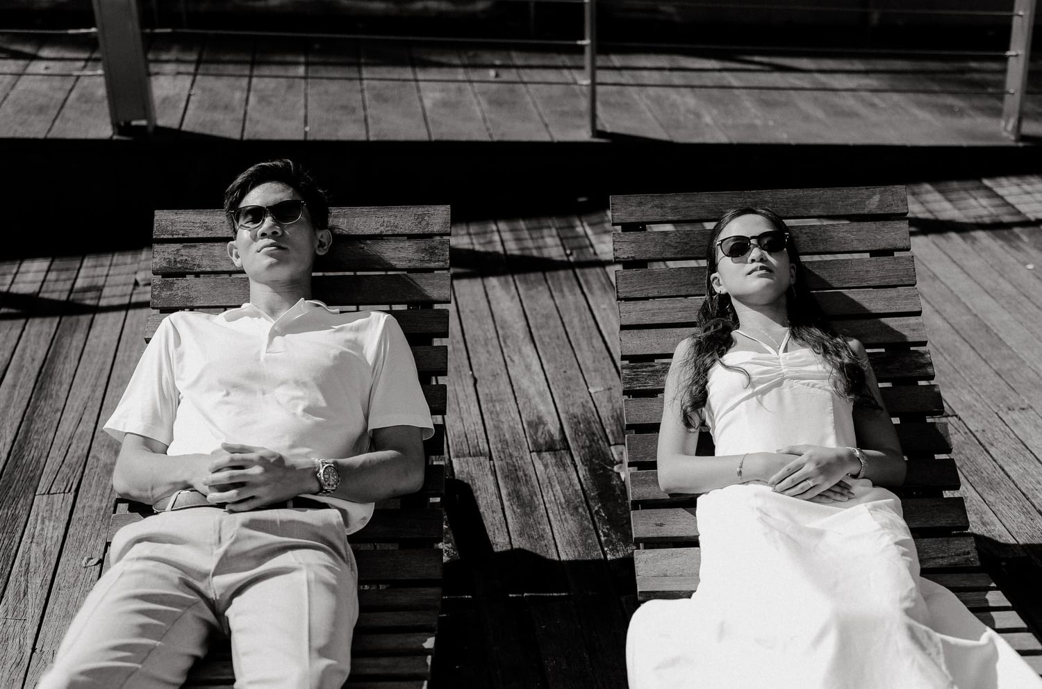 A couple relaxes on wooden decking in black and white, wearing sunglasses and matching outfits.