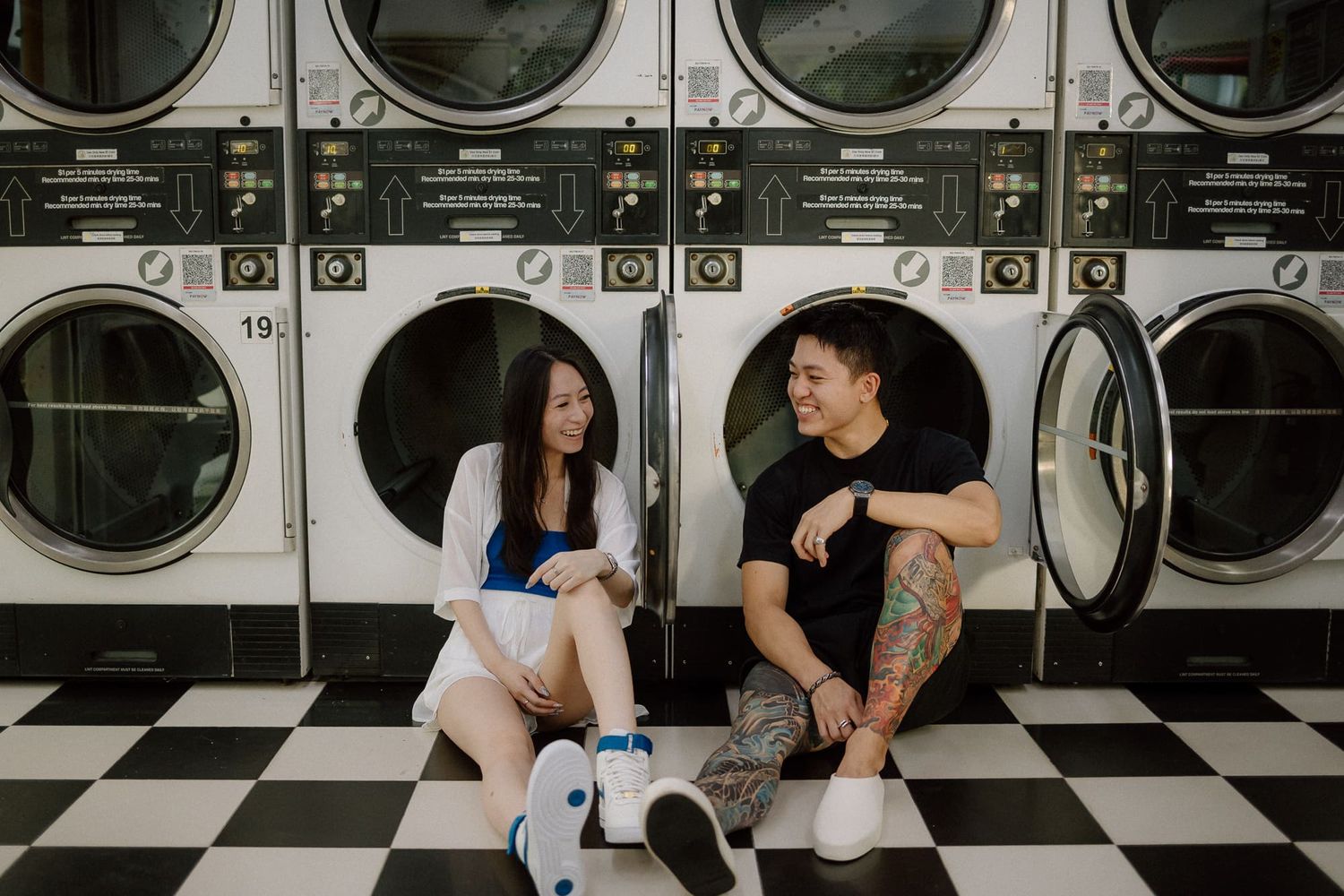 Two people sit together and laugh while doing laundry at a retro-style laundromat with checkered floors.