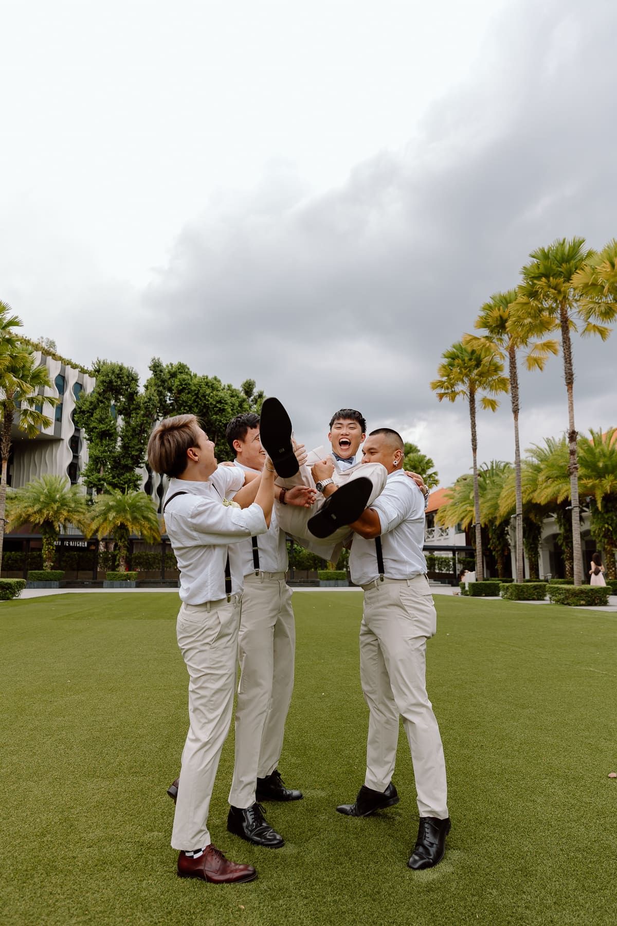Groomsmen in light colored suits playfully lifting someone in celebration on grass lawn.