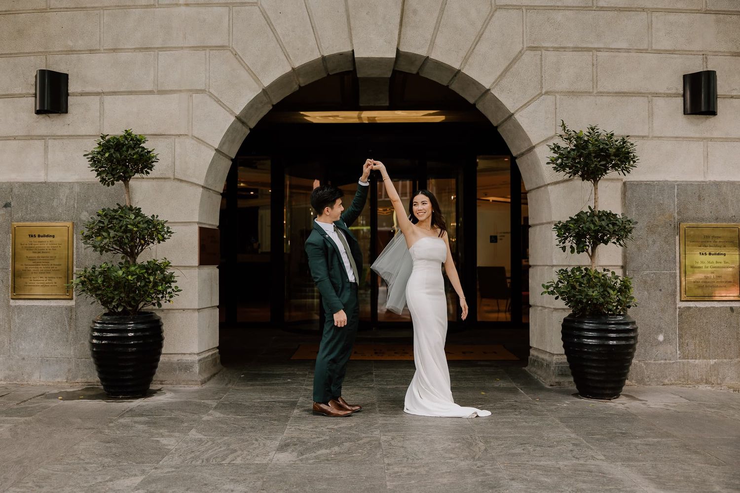 A couple celebrates their wedding day in front of an elegant arched entrance with potted plants.