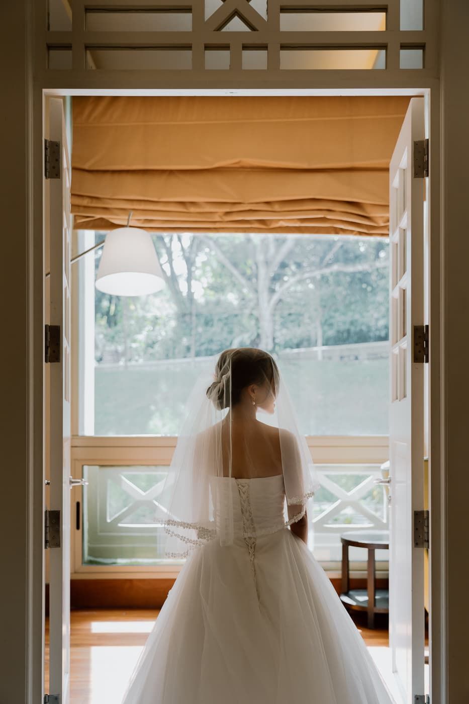 Bride in flowing wedding gown stands silhouetted against window with orange roman shade.