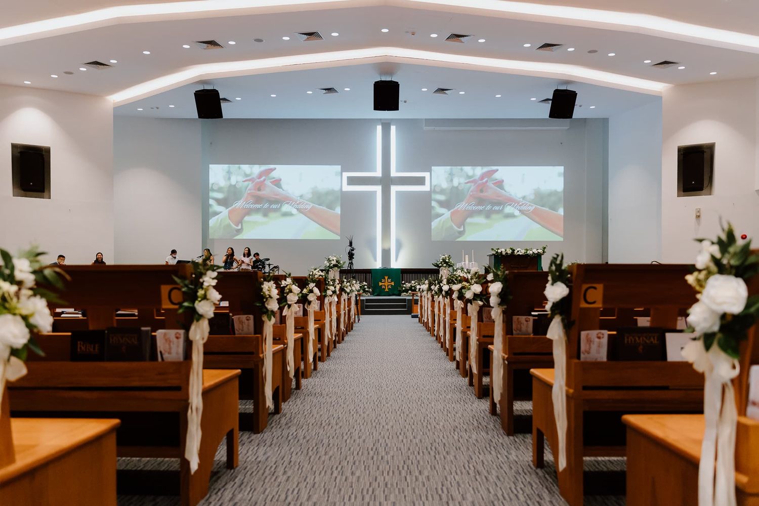Modern church sanctuary with illuminated cross and dual projection screens decorated with white floral arrangements.