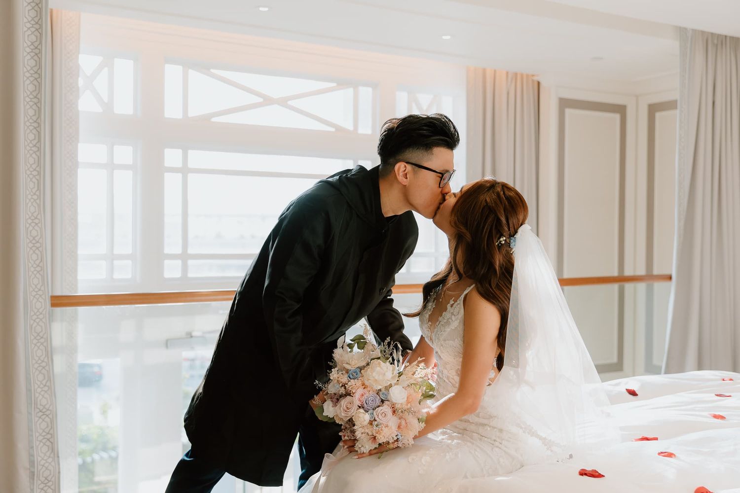 Romantic wedding couple poses with pink bouquet in bright sunlit room.