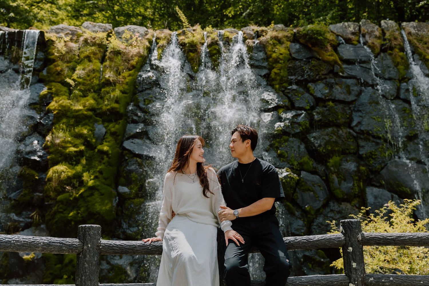 Couple holding hands in front of a mossy stone waterfall surrounded by lush green trees.