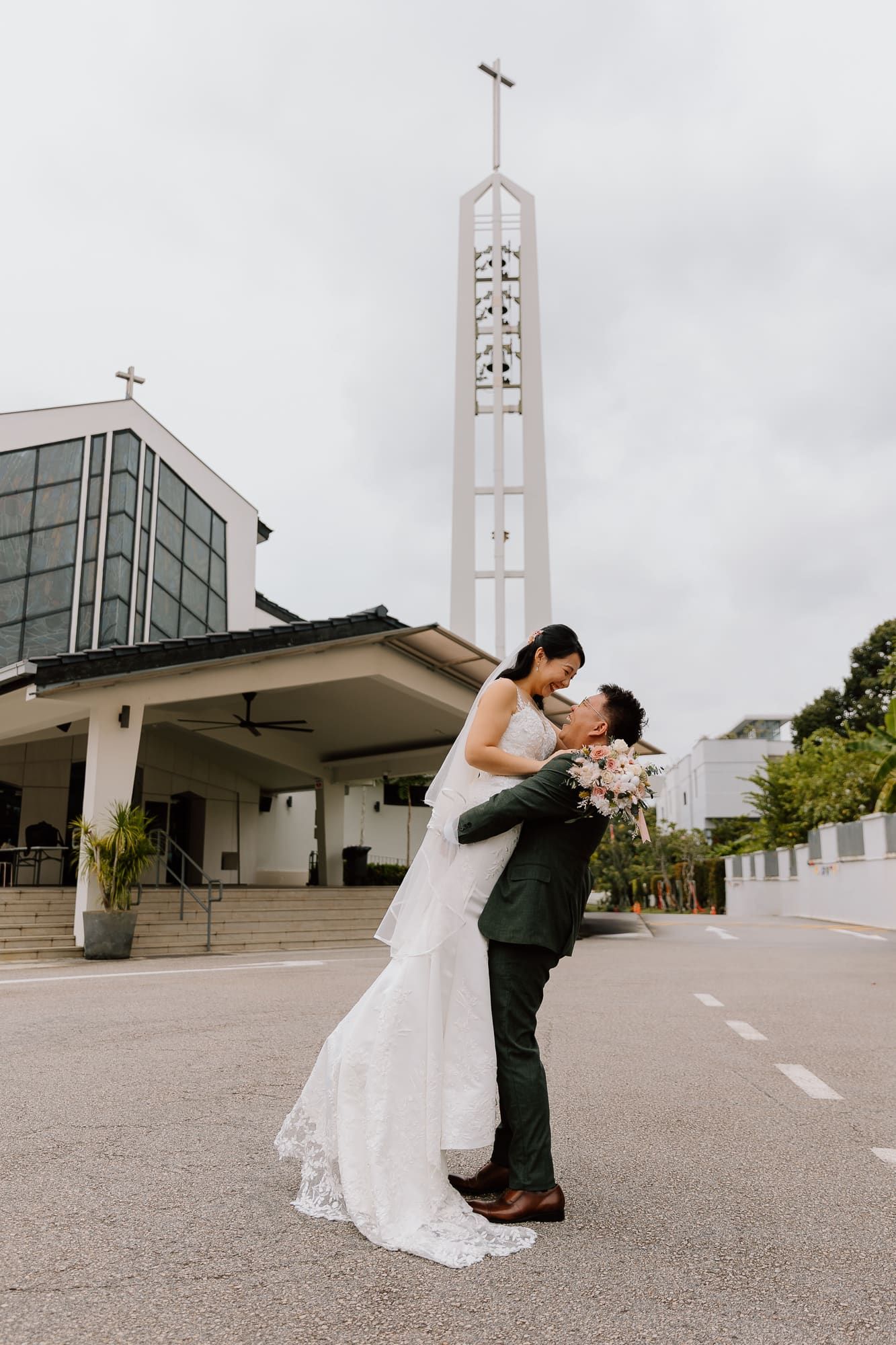 A couple shares a romantic dip kiss outside a modern church with a tall spire against an overcast sky.