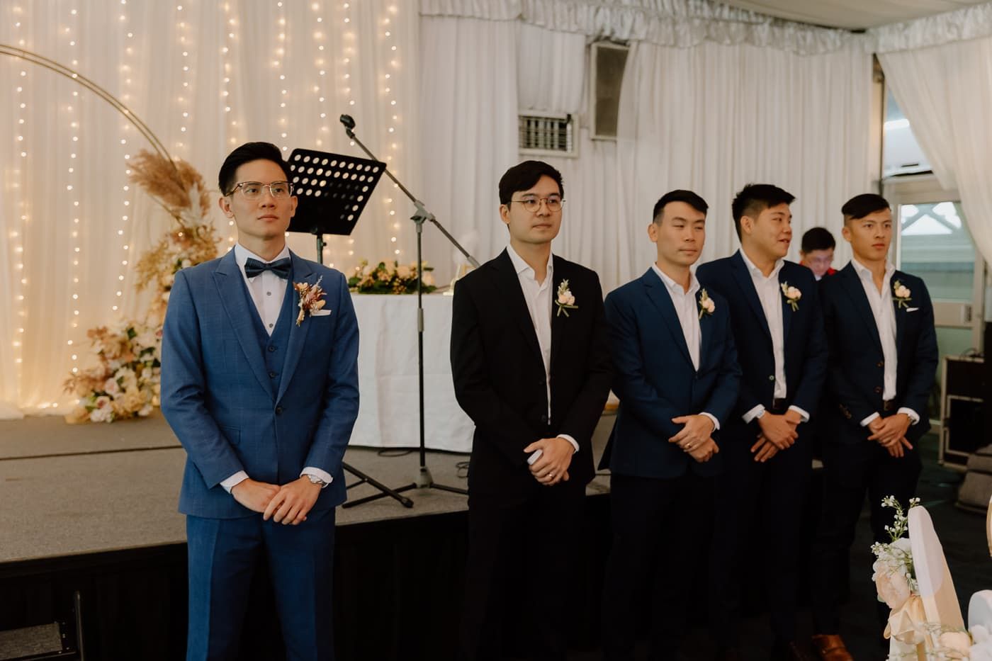 Groomsmen line up at wedding ceremony wearing coordinated navy suits and boutonnieres.