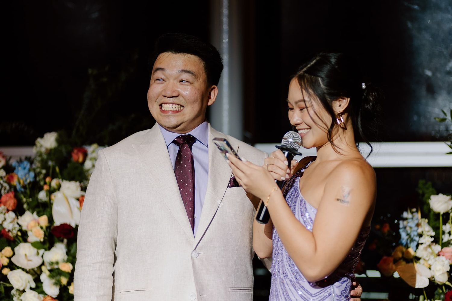 Wedding guests share microphone during reception speeches with floral backdrop.