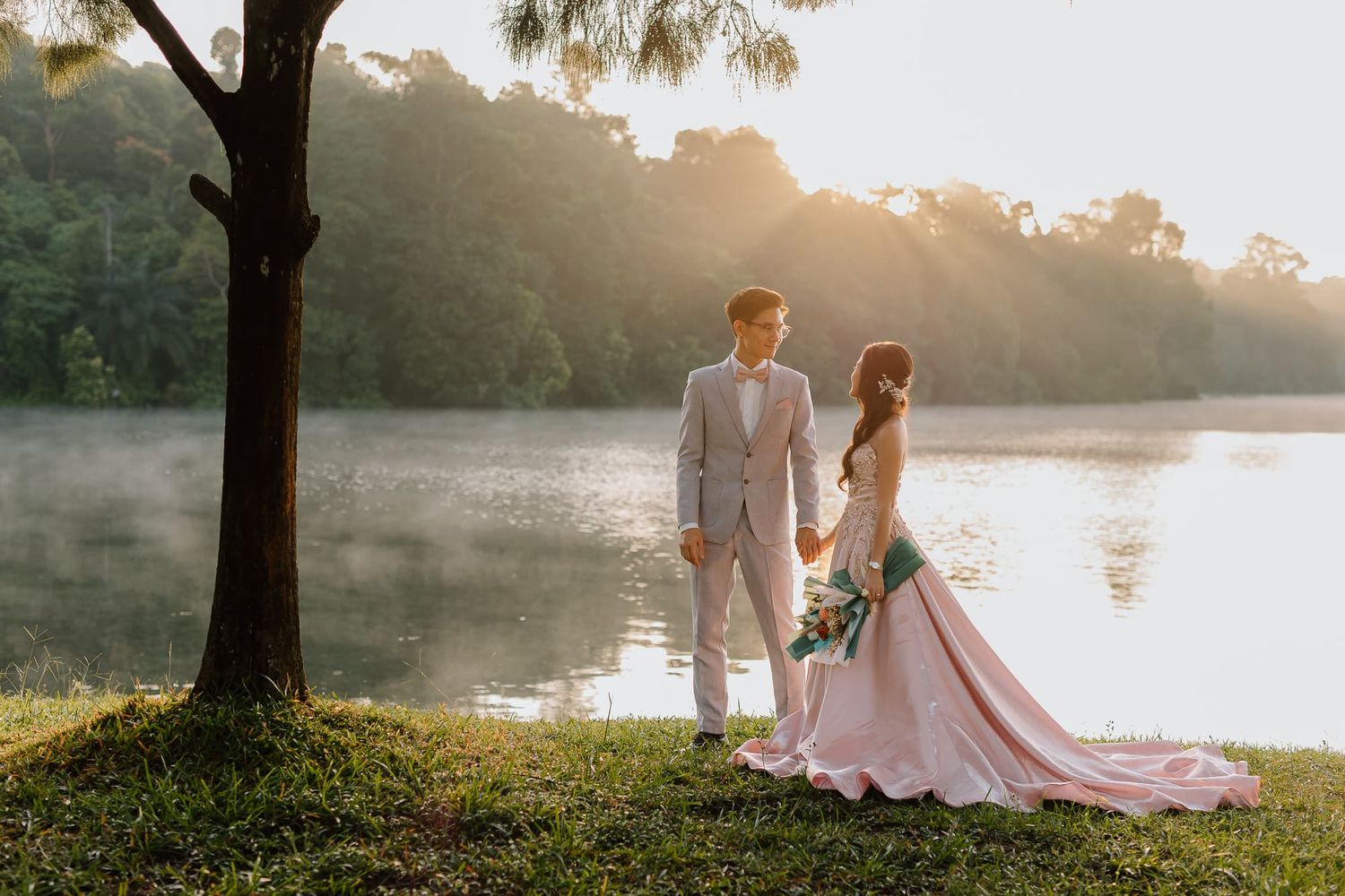 Couple shares tender moment under tree branches beside peaceful lake at sunset.