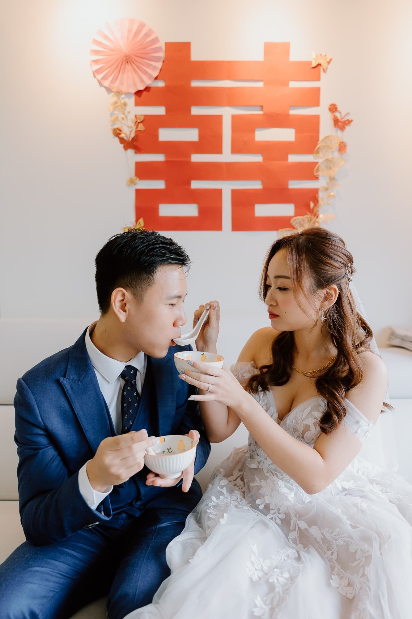 Wedding couple playfully feeding each other in front of red double happiness symbol.