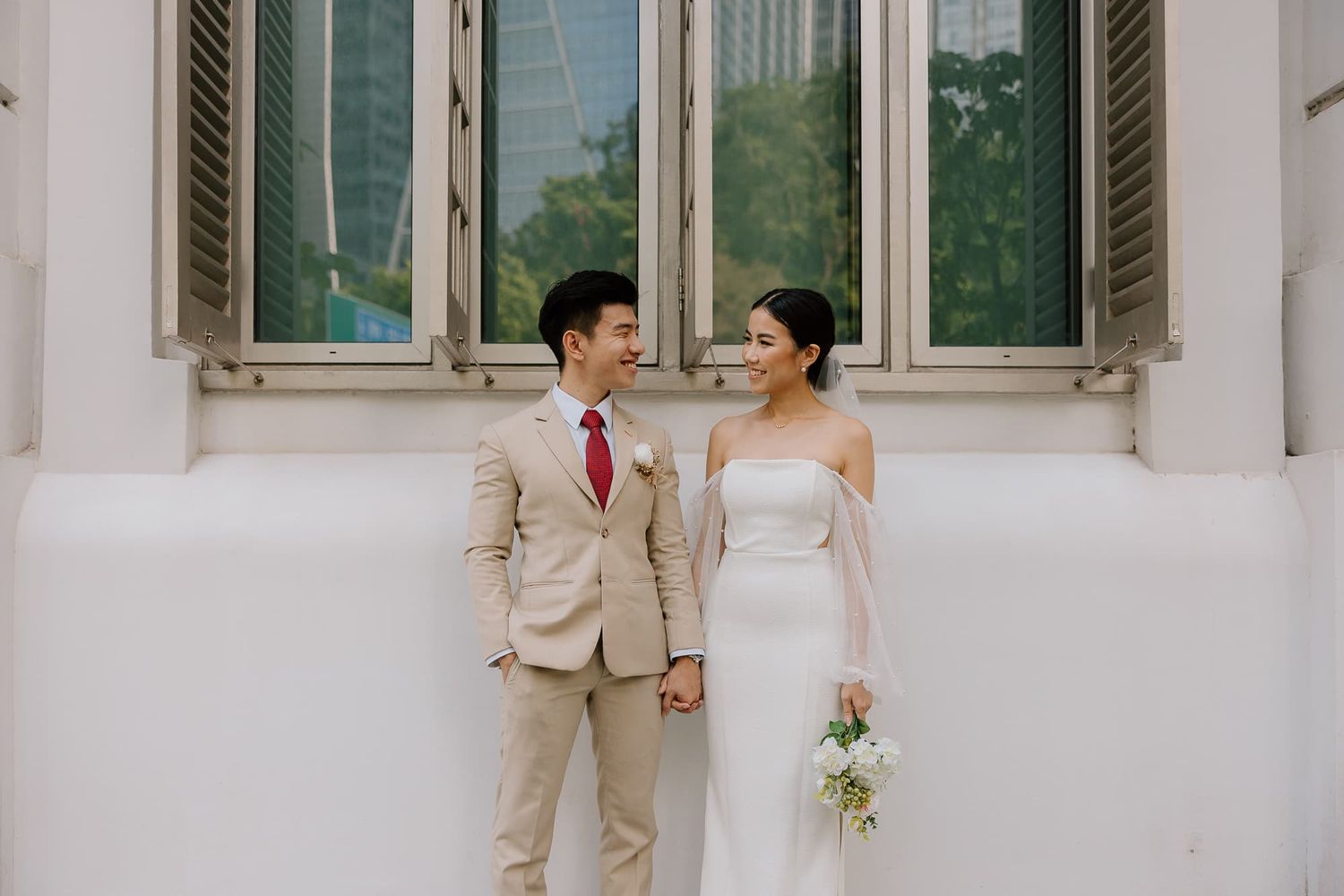 A couple in wedding attire poses against a white building with shuttered windows.