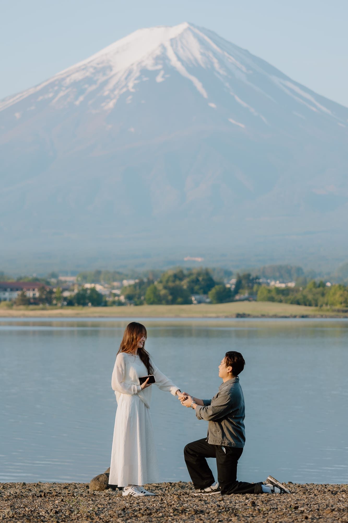 Man proposes to woman on one knee by a serene lake with iconic snow-capped Mount Fuji in the background.