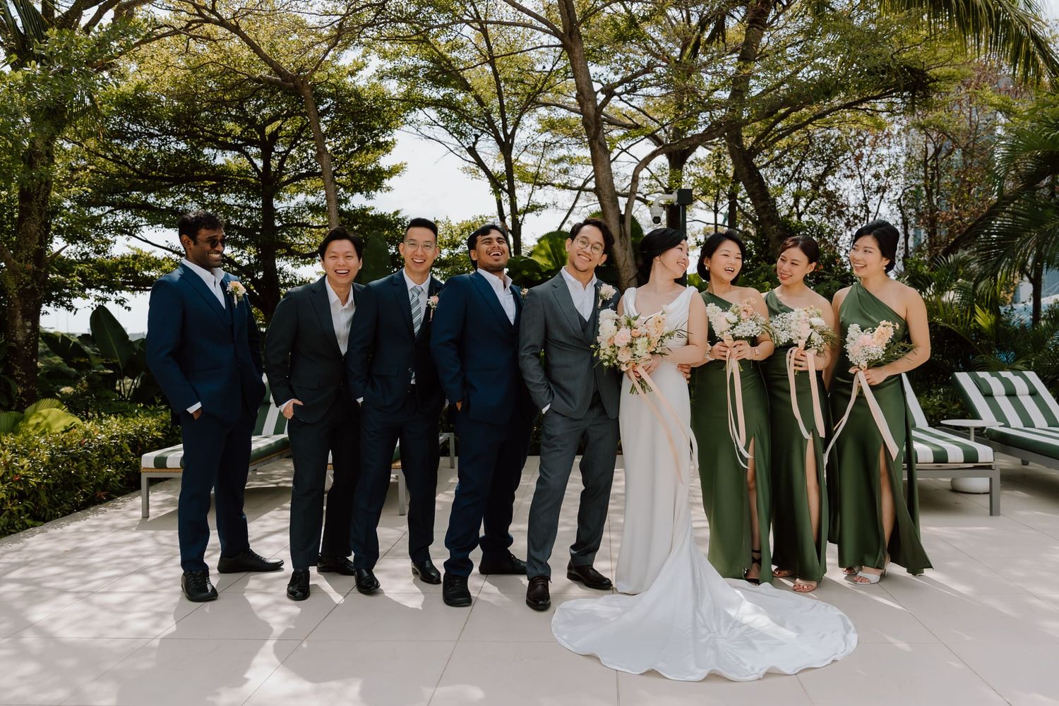Wedding party in formal attire poses outdoors with palm trees and greenery in the background.