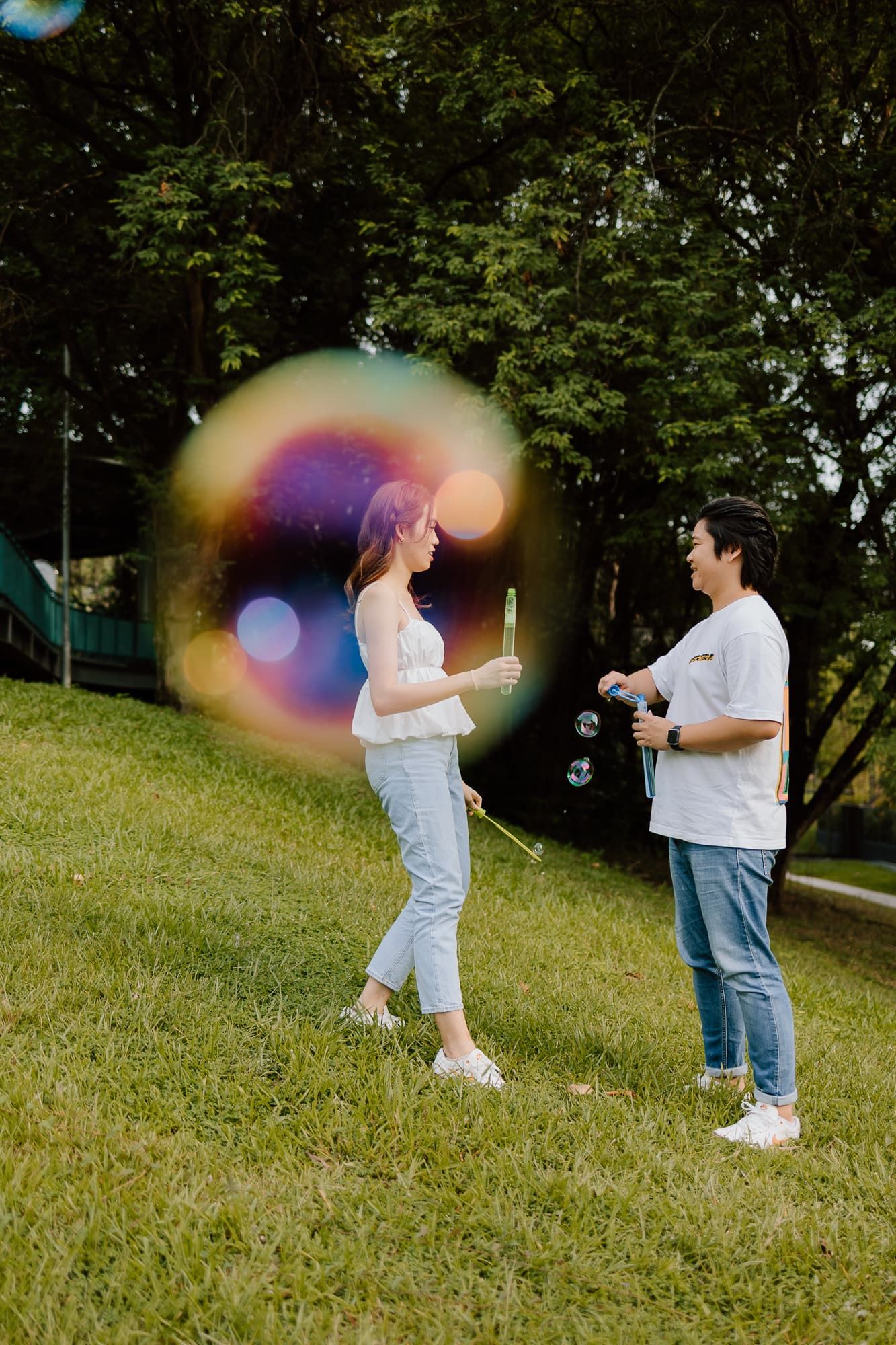Couple playing with soap bubbles creating rainbow effect in park.
