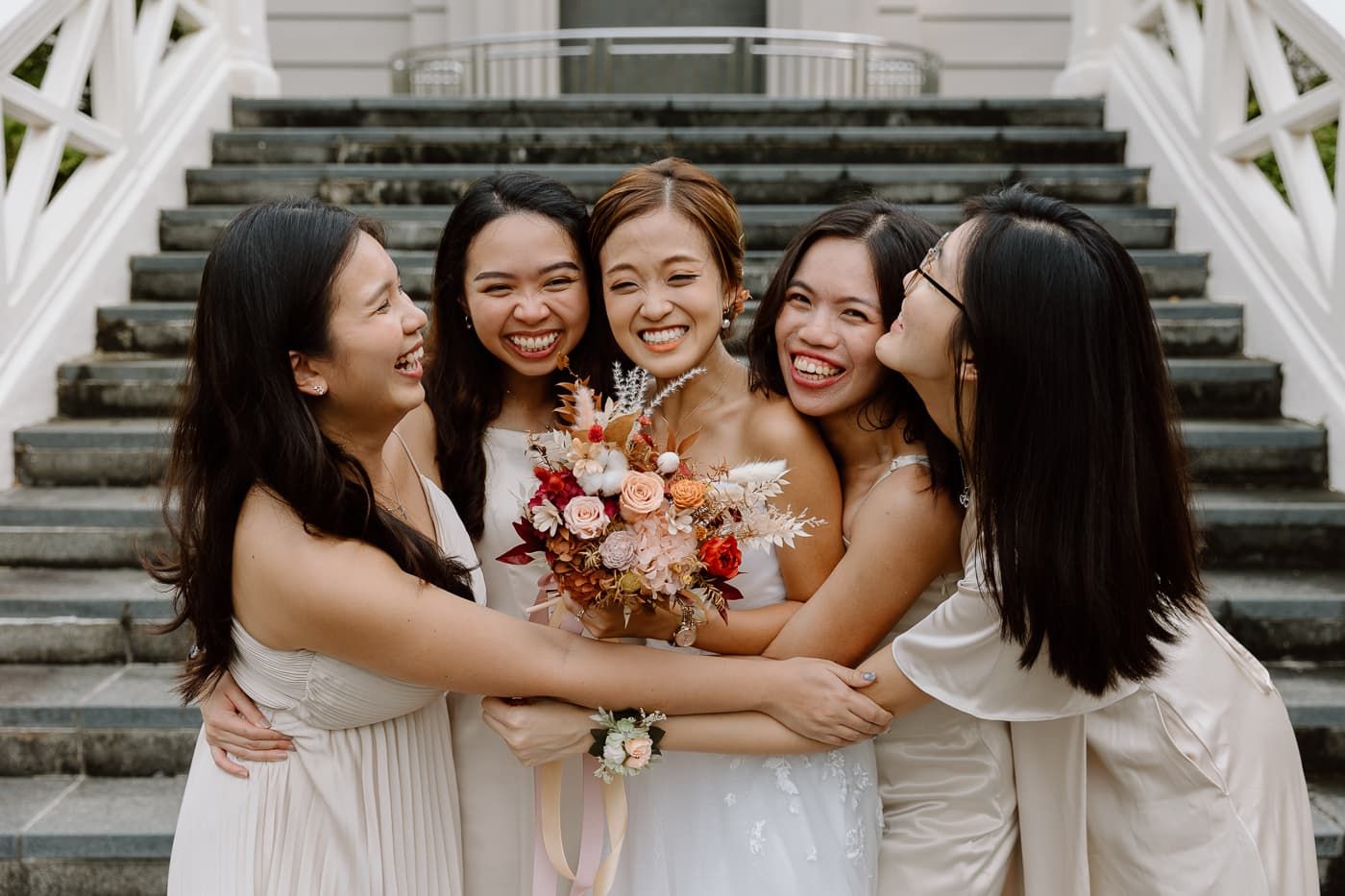 Bridesmaids in white dresses sharing candid laughter on outdoor steps.
