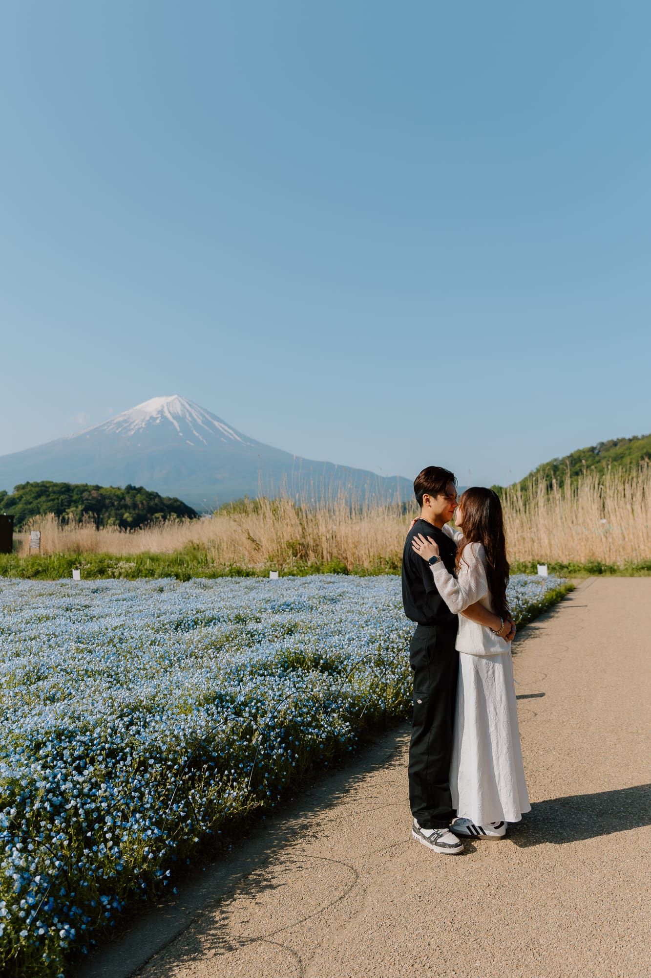 Couple embracing in blue flower field with Mount Fuji in background under clear sky in Japan.