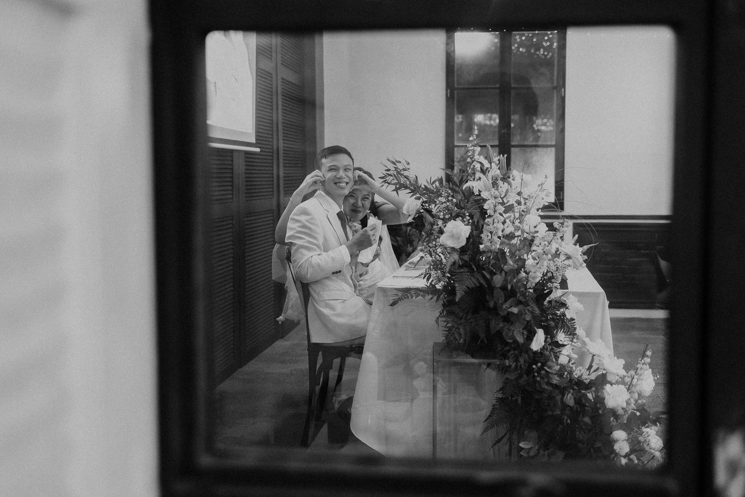 A person in a long coat and hat smells flowers at a vintage sidewalk flower stand in black and white.