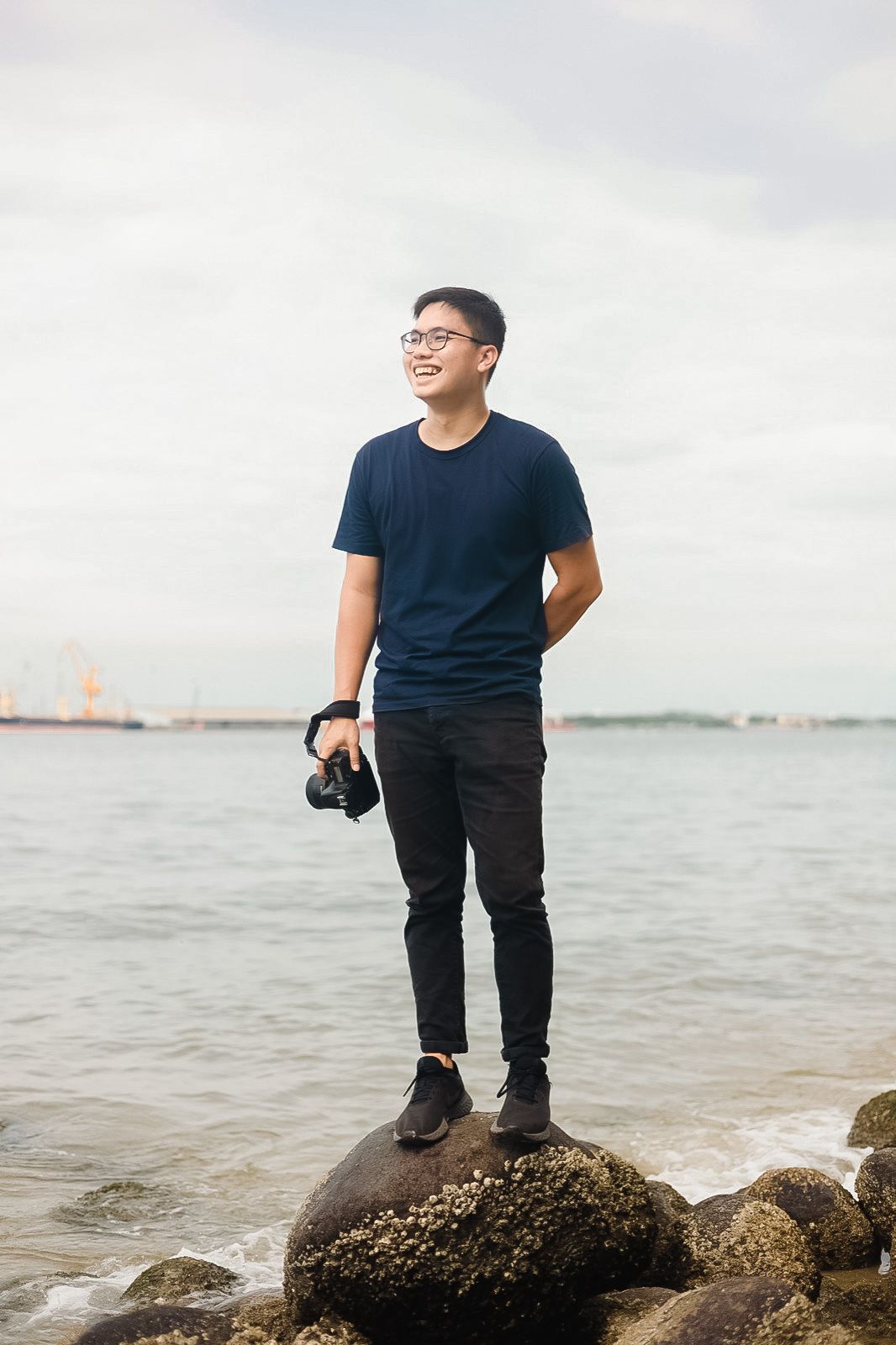 Photographer stands on coastal rocks holding camera gear against a cloudy harbor backdrop.