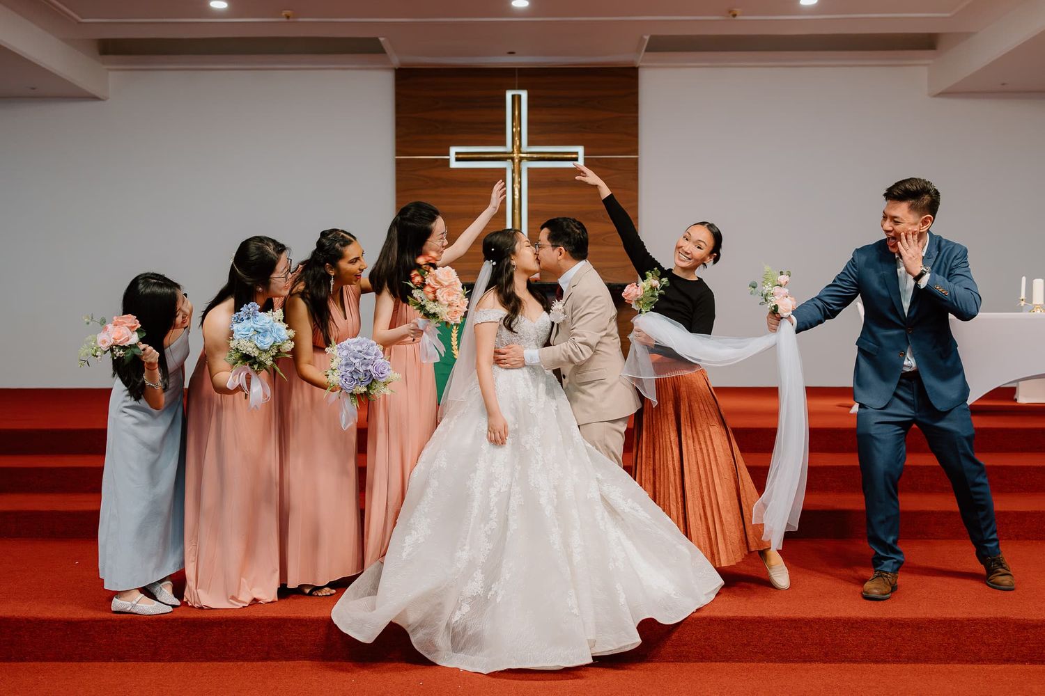 Bridal party poses playfully on church stage with bridesmaids in pink dresses.