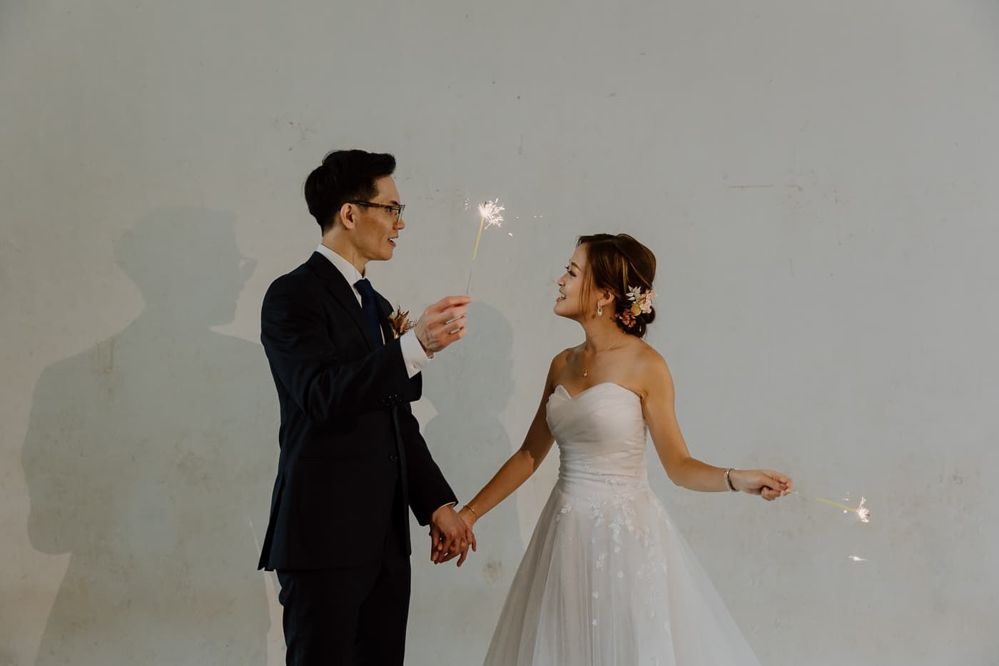 Couple shares romantic sparkler moment against white wall during wedding celebration.