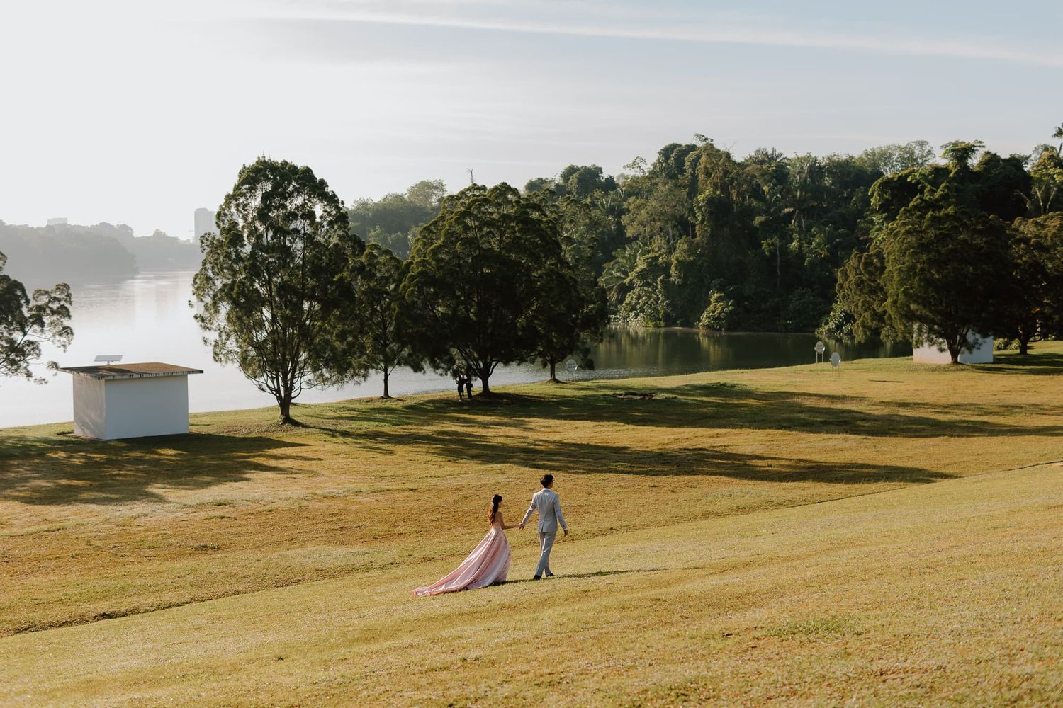 Distant view of couple walking together on grassy hill with trees and lake in background.