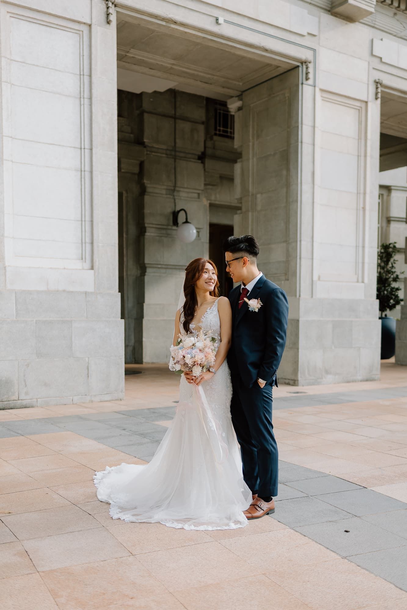 Wedding couple poses romantically against marble columns in a grand building entrance.
