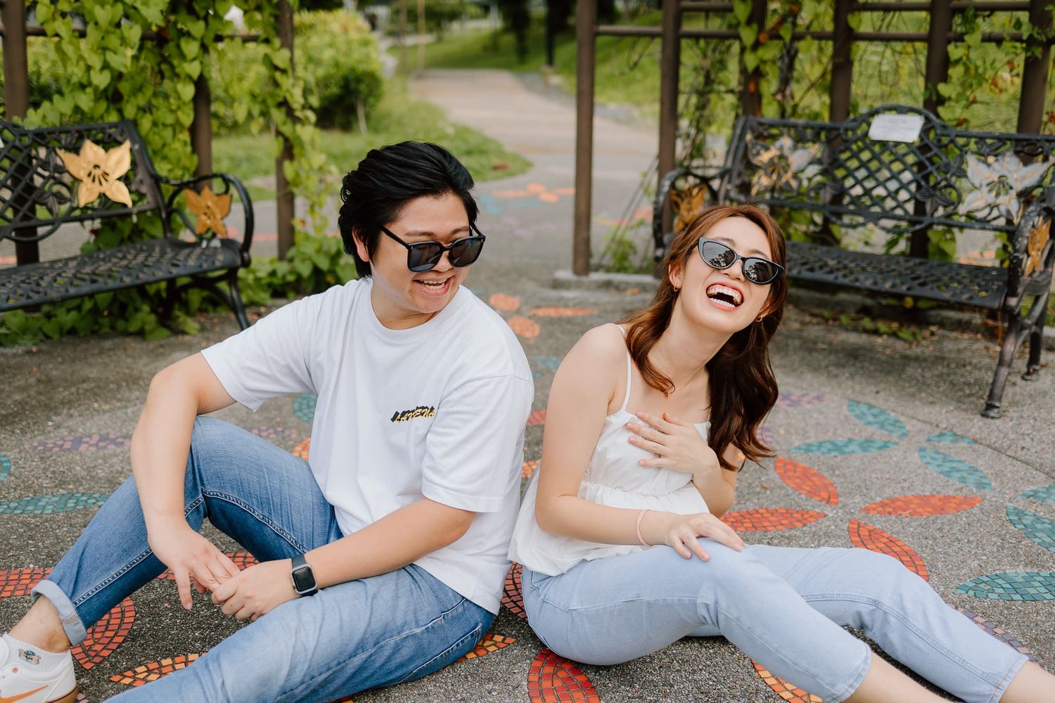 Couple sitting on ground sharing joyful laughter in outdoor garden setting wearing casual attire.