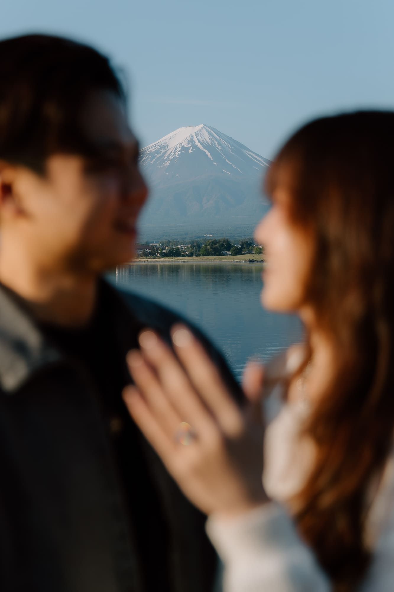 Couple gazing at each other with snow-capped Mount Fuji reflected in lake behind them, engagement ring visible.
