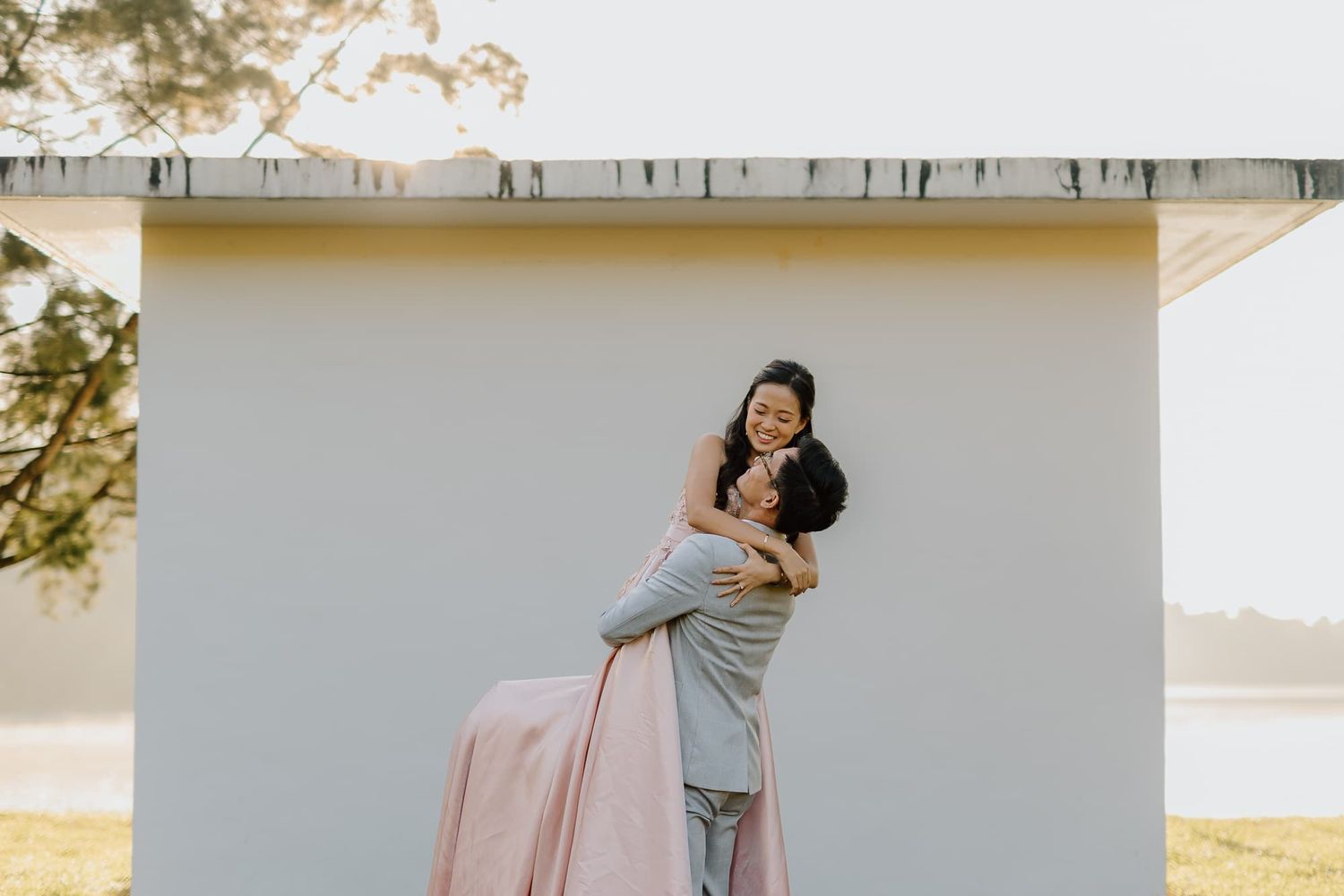 Playful couple embracing against a white wall with water and nature in the background.