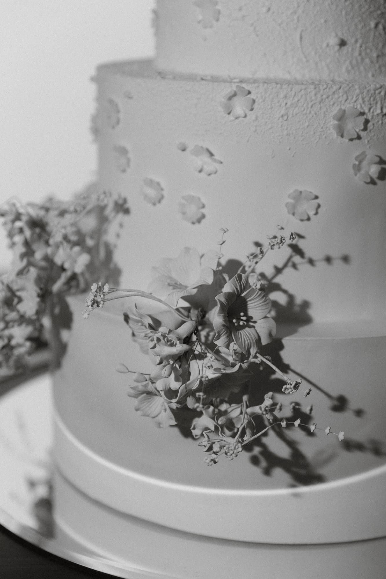 Close-up black and white photo of delicate floral decorations on the edge of an elegant white wedding cake.