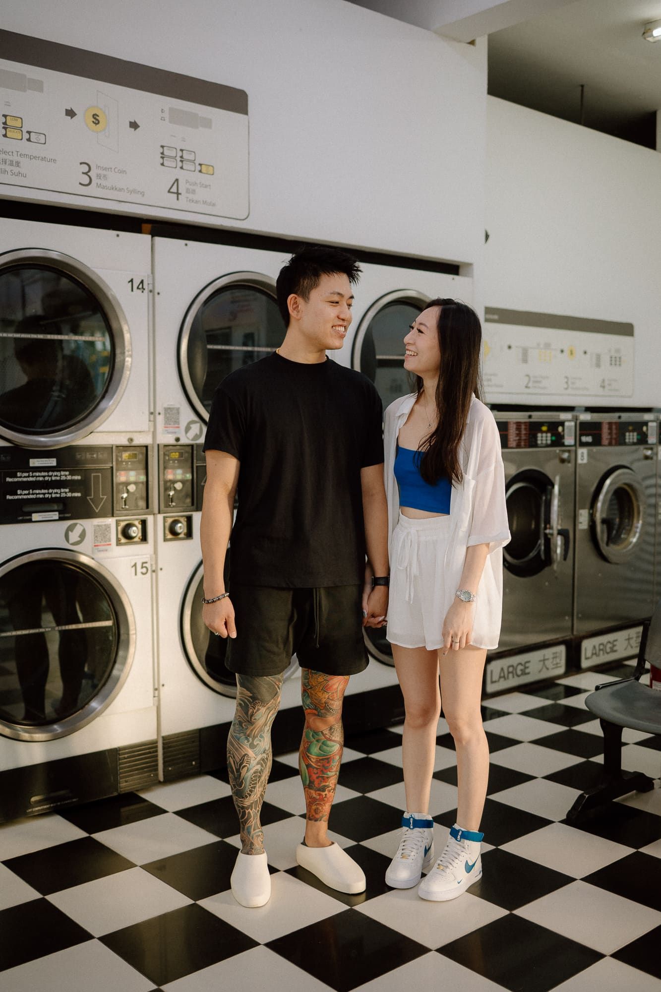 A couple stands together in a laundromat with black and white checkered floors and stacked washing machines.