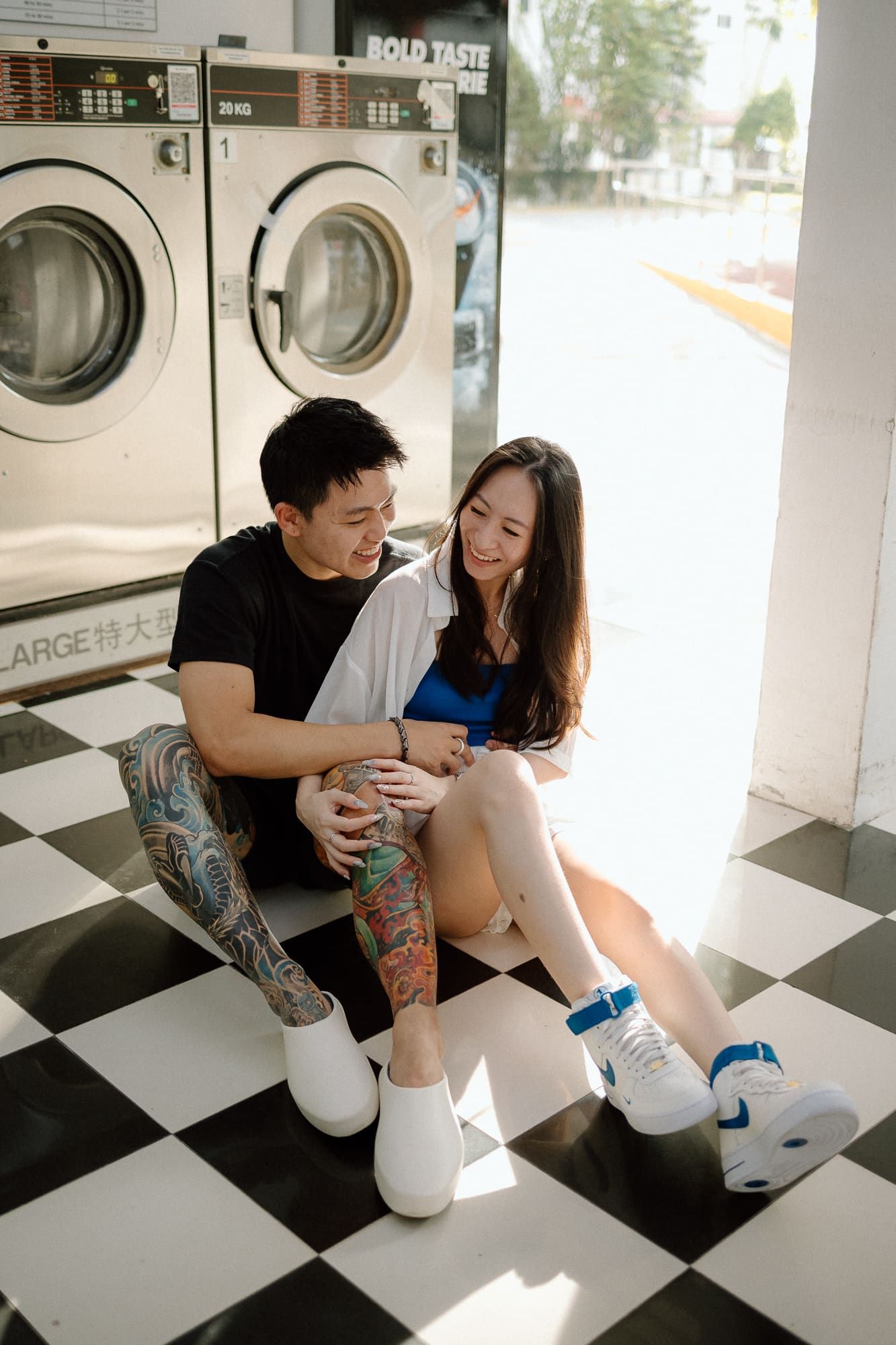 A couple shares an intimate moment while sitting on the floor of a modern laundromat with black and white checkered tiles.