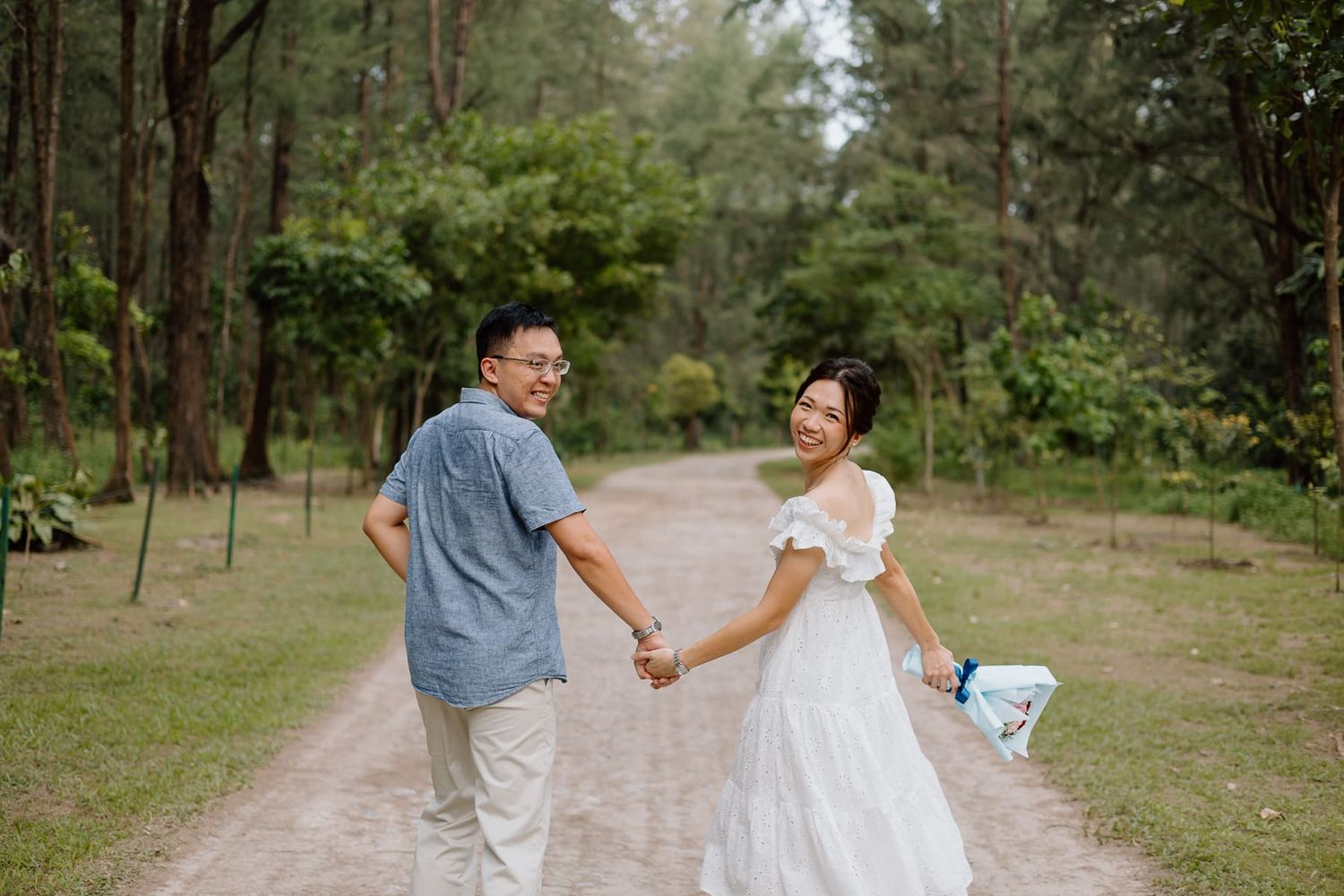 A couple walks hand in hand down a sandy nature path surrounded by pine trees.