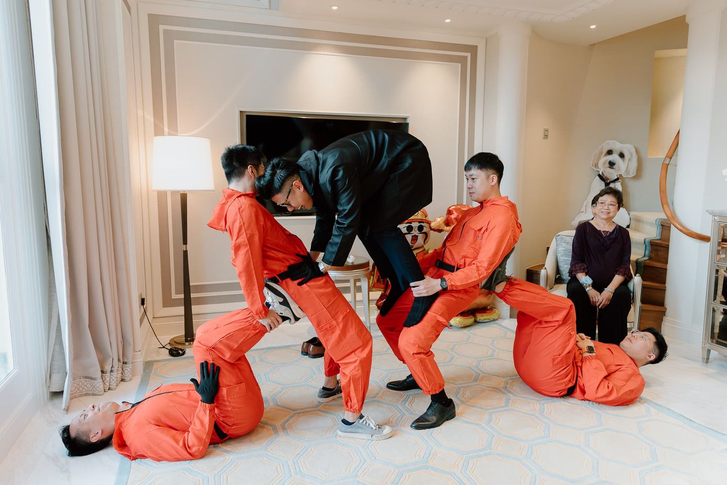 Wedding party members in bright orange jumpsuits and formal wear pose in sequence across a white room.